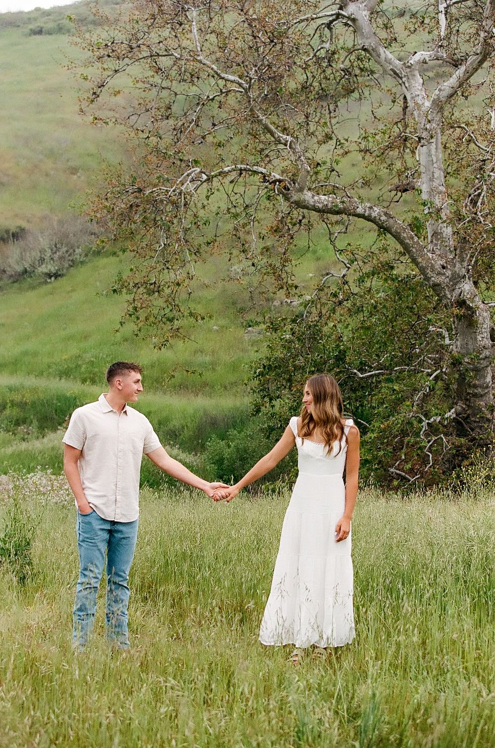 Couple holding hands in a lush green meadow beneath a large oak tree, woman in white dress.