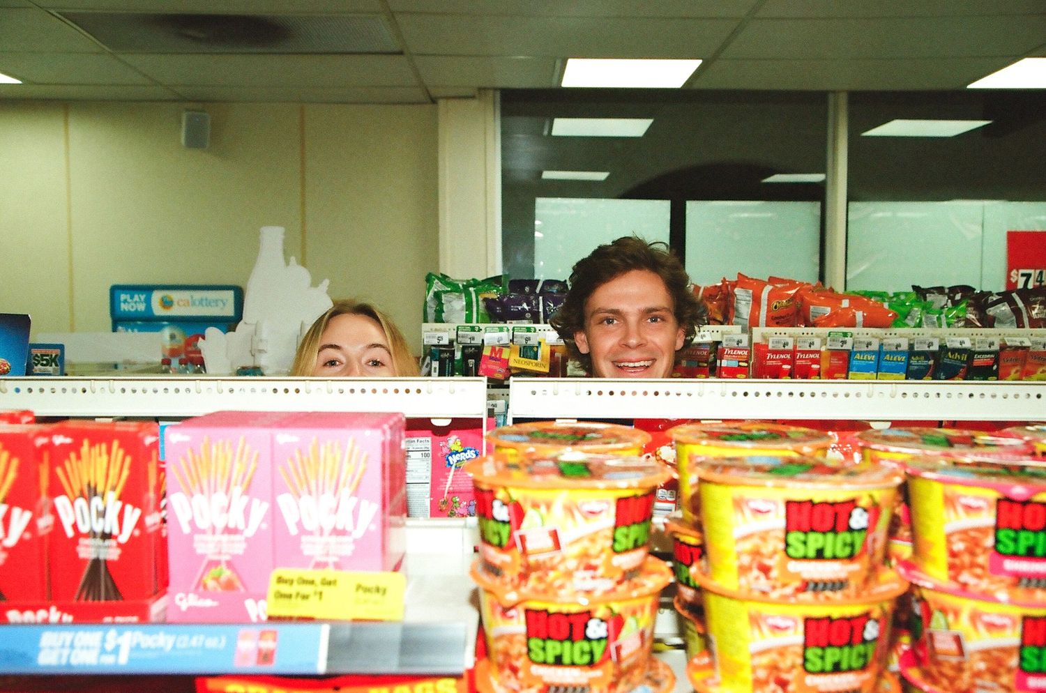 Smiling couple peeking over a convenience store shelf stocked with Pocky and Hot Spicy snacks.