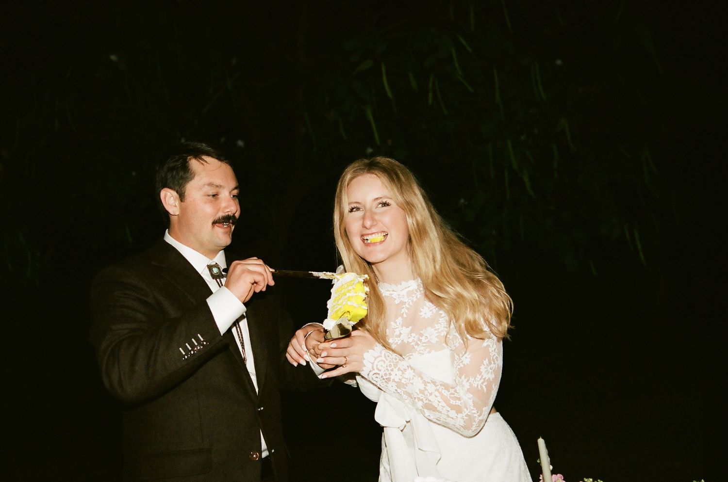 Couple in formal attire sharing a drink at a nighttime wedding reception, bride in white lace gown.