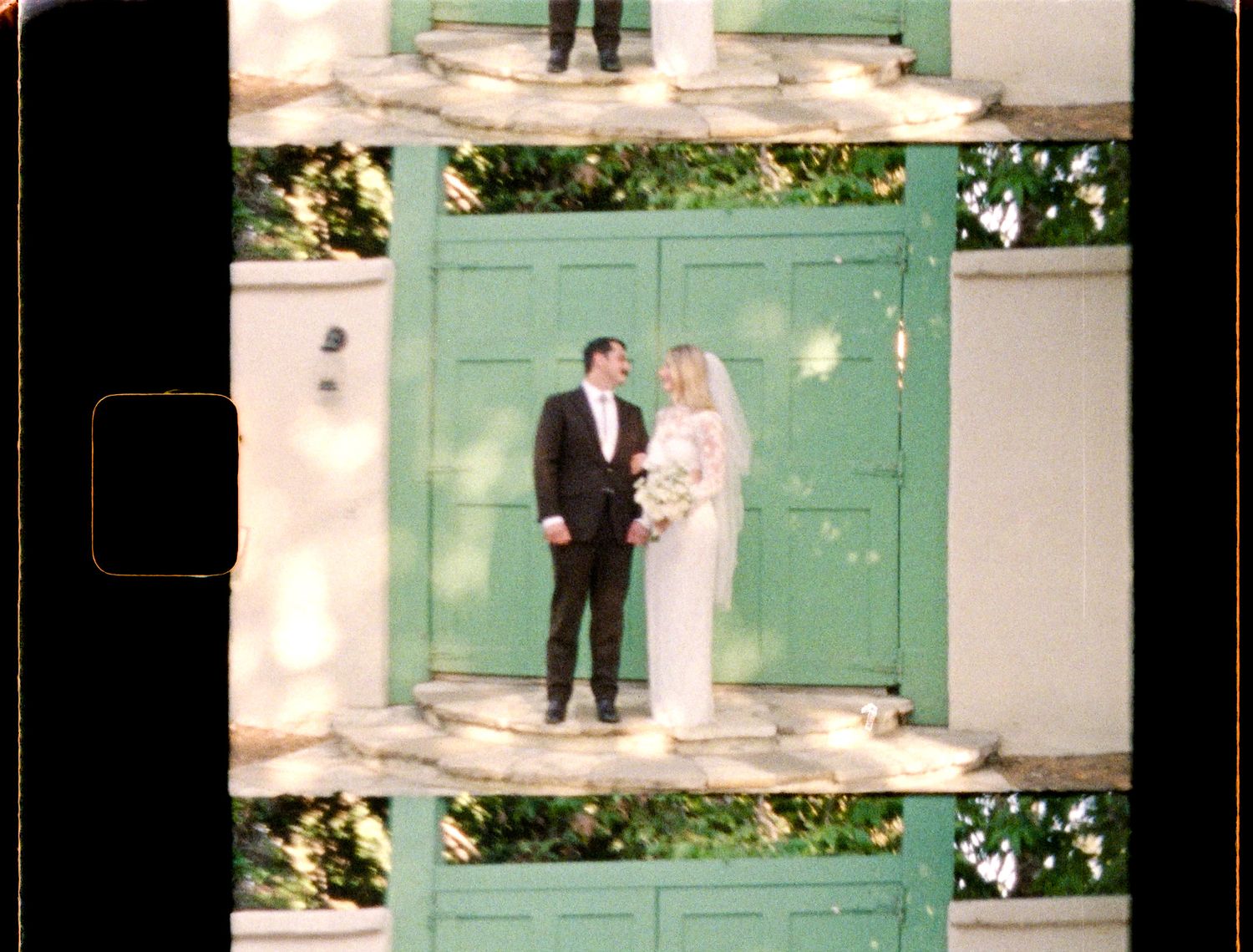 Film photo of a bride and groom standing before green double doors, surrounded by lush greenery on their wedding day.