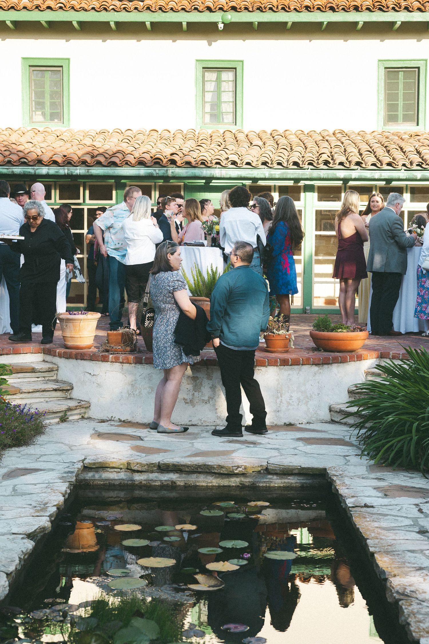 Guests mingle at an elegant outdoor courtyard event near a lily pond at a Spanish-style venue.