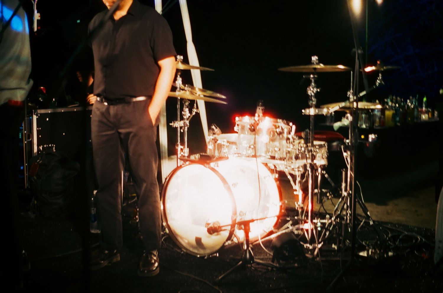 Drummer standing behind a glowing white bass drum kit on a dark outdoor stage during a live music performance.