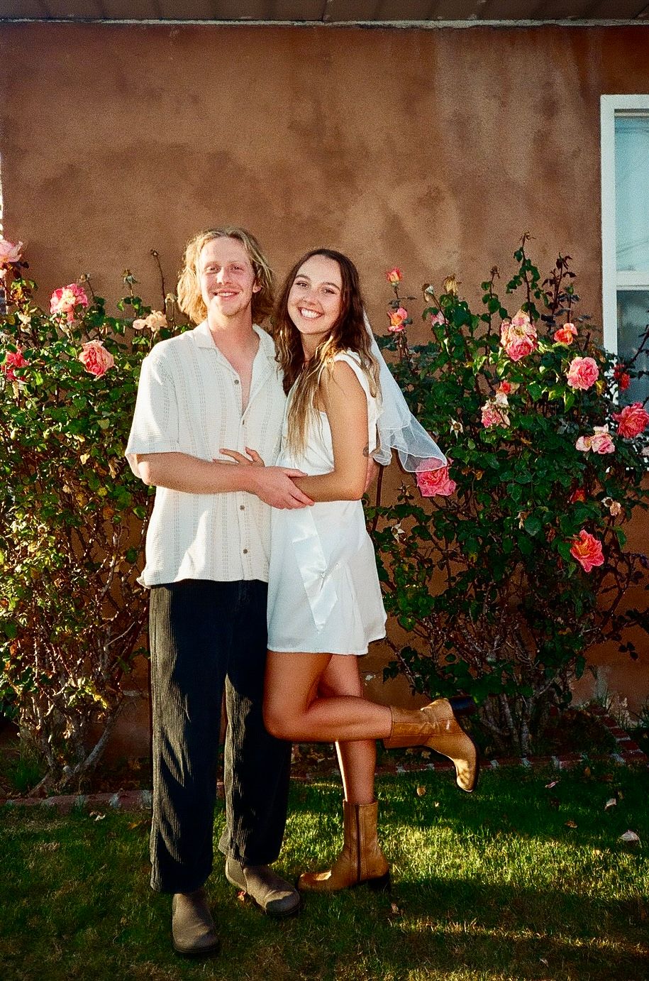 Smiling couple posing in front of blooming pink roses outside an adobe wall on a sunny day.