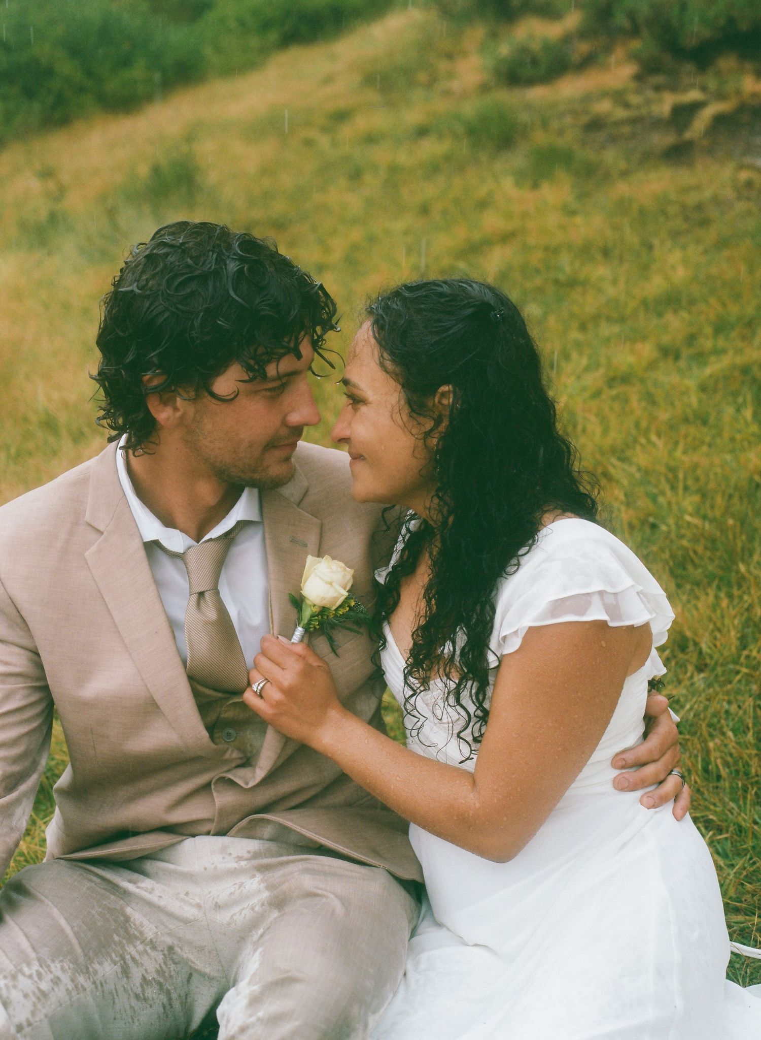 A couple in wedding attire sits on a grassy hillside, sharing a romantic moment with a white rose boutonniere.