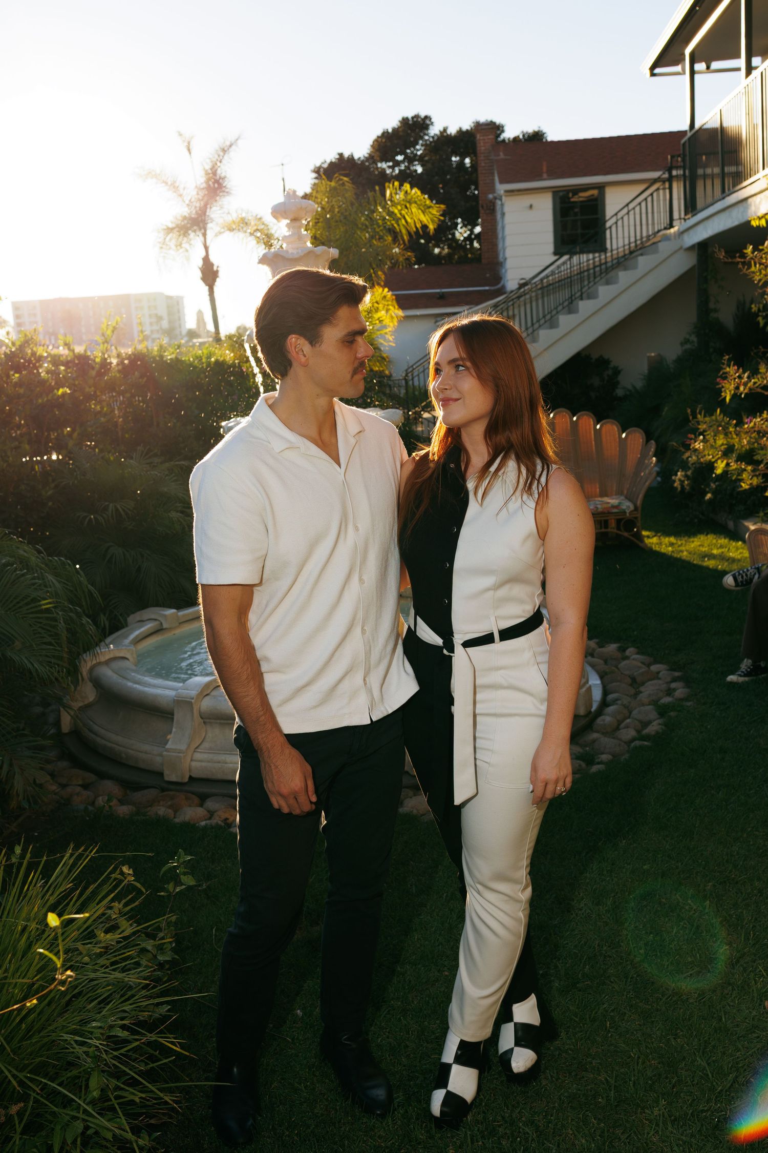 Couple standing in sunny garden by fountain, man in white shirt and woman in black and white outfit.