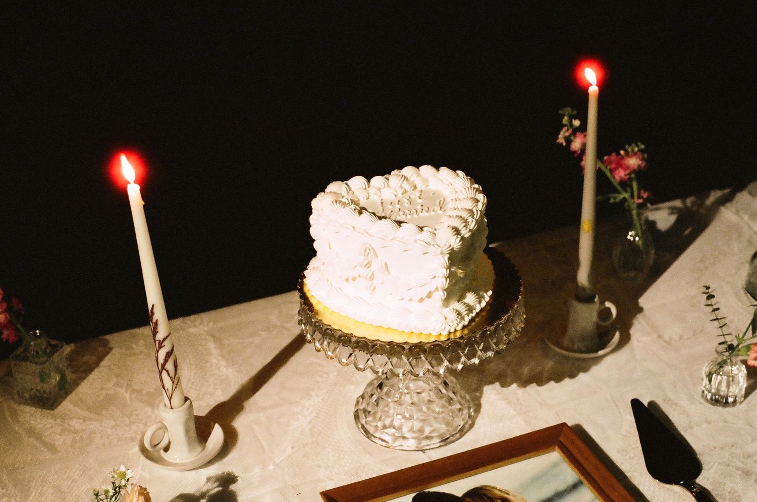 White frosted wedding cake on crystal stand flanked by two lit red taper candles on a decorated table.