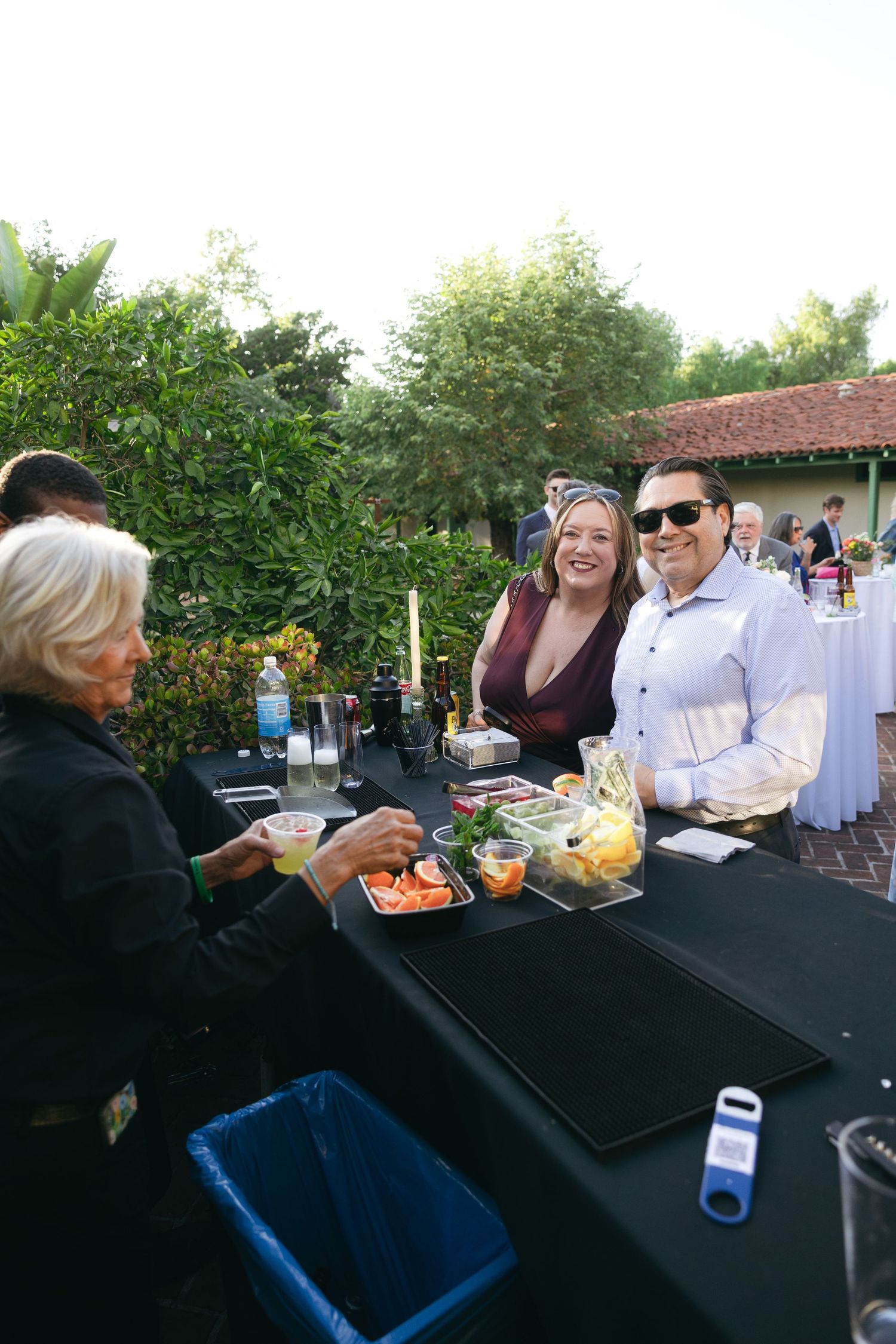 Guests smile at an outdoor catered event with food trays on a black-draped table surrounded by lush greenery.