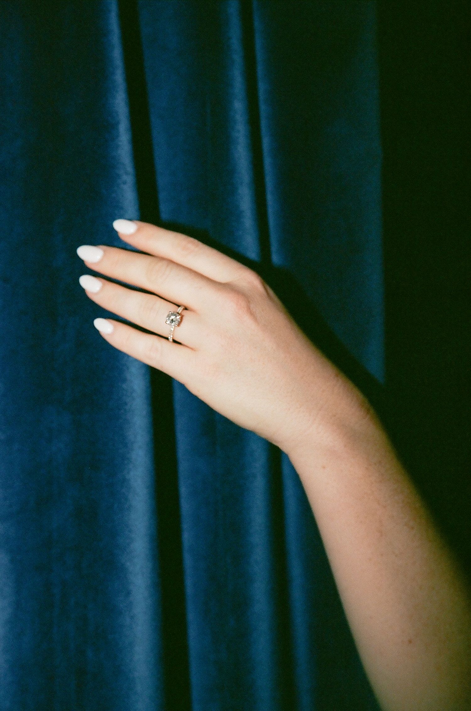 Close-up of hand wearing a delicate gold solitaire diamond engagement ring against blue velvet.