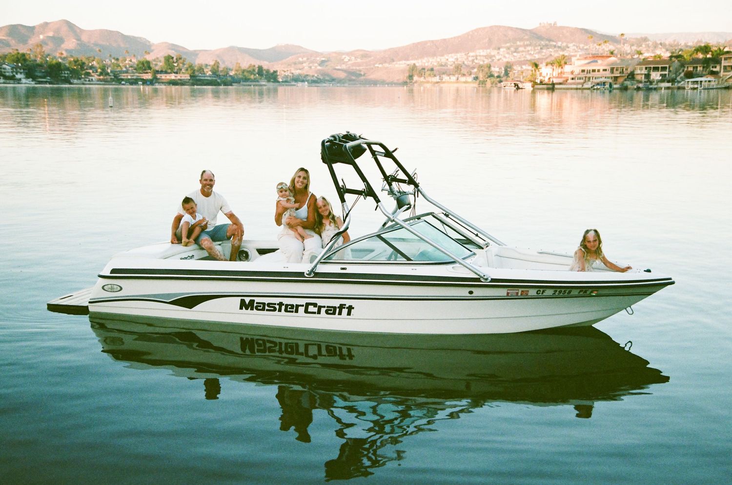 A family relaxes on a MasterCraft boat on a calm lake with a scenic hillside town in the background.