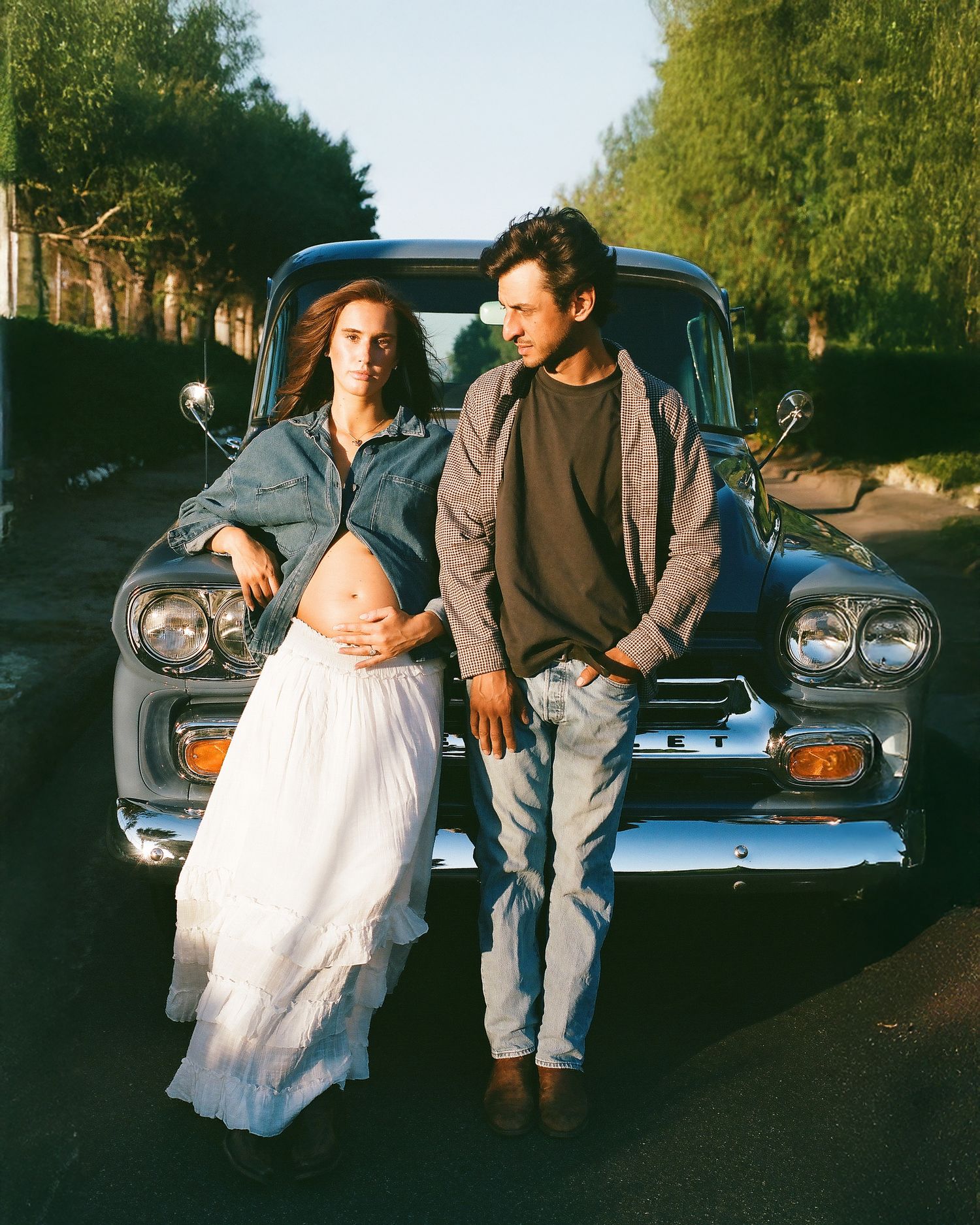 Expecting couple stands before vintage blue truck, man touching womans baby bump at golden hour.