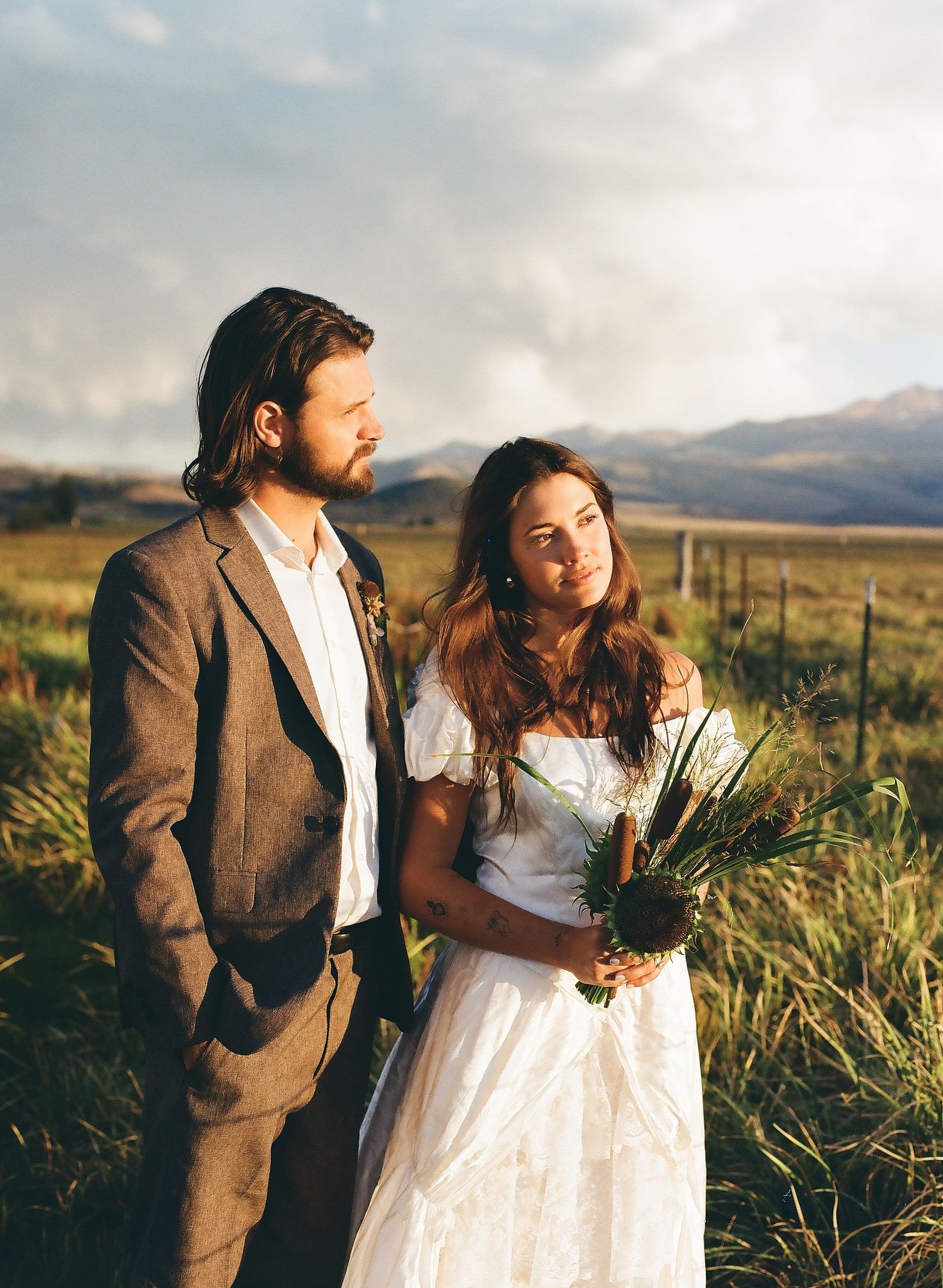 Bohemian bride and groom stand in a golden meadow with mountain backdrop, bride holding wildflower bouquet.