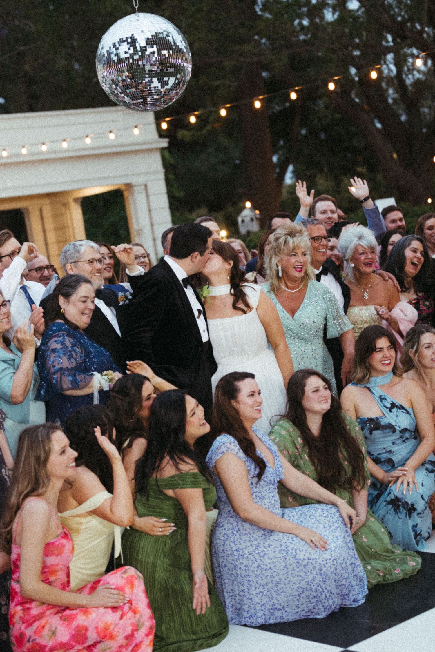 Bride and groom share a kiss surrounded by cheering wedding guests under a disco ball at an outdoor reception.