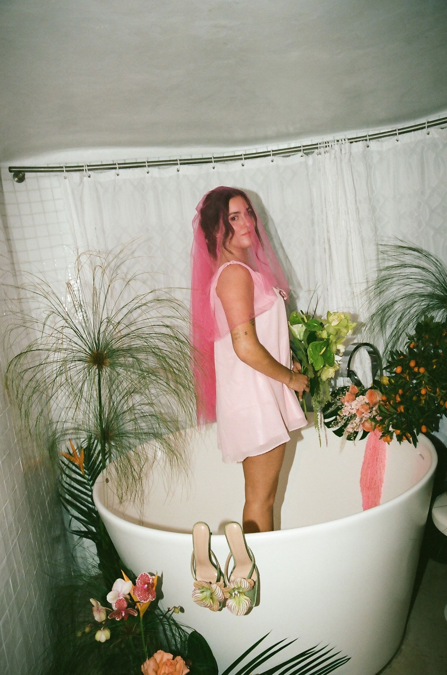 Bride in pink slip dress with pink veil standing in white bathtub surrounded by tropical flowers.