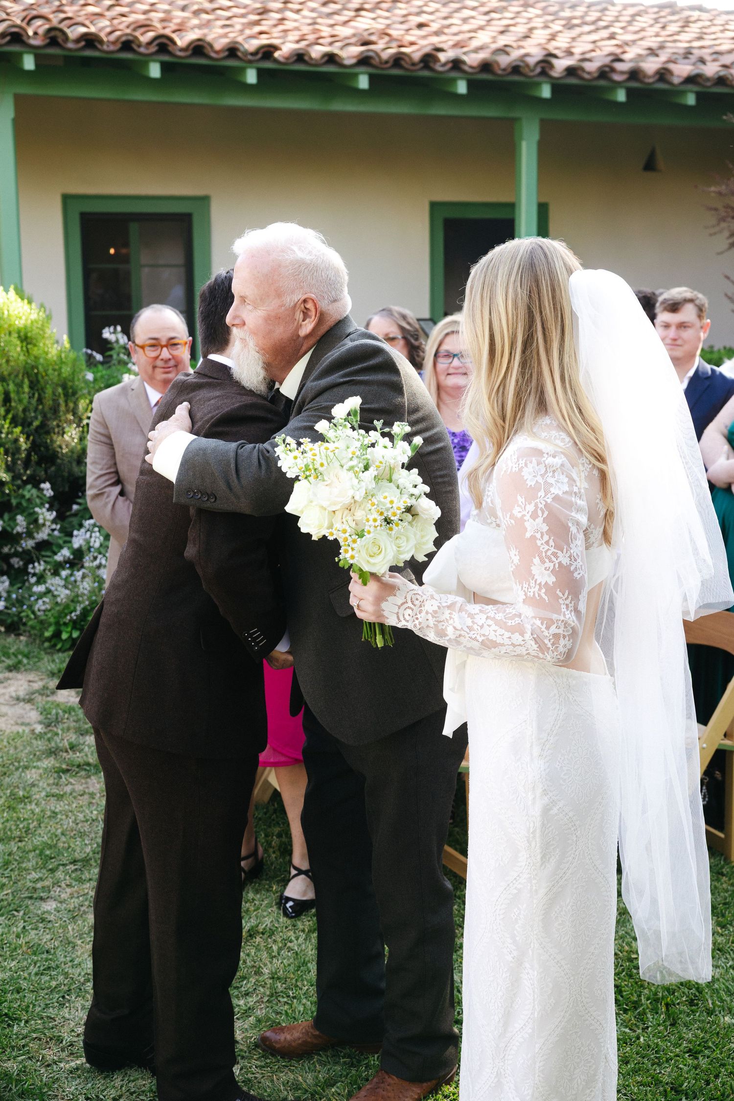 Bride in lace gown holds bouquet as older man shares emotional hug during outdoor wedding ceremony.