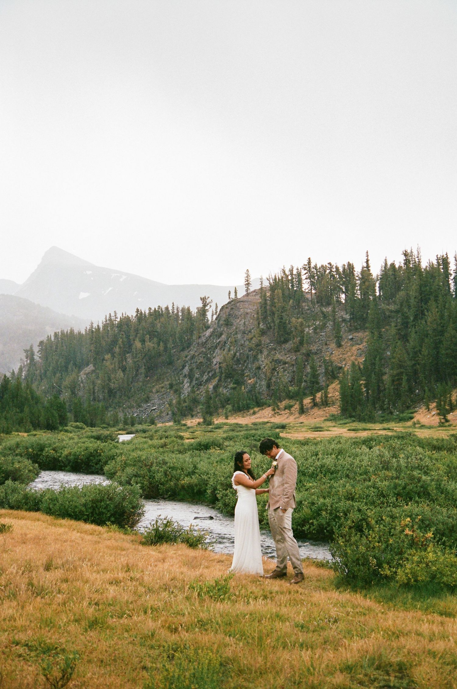 Couple dancing in white dress and tan suit by a mountain stream surrounded by evergreen trees and misty peaks.