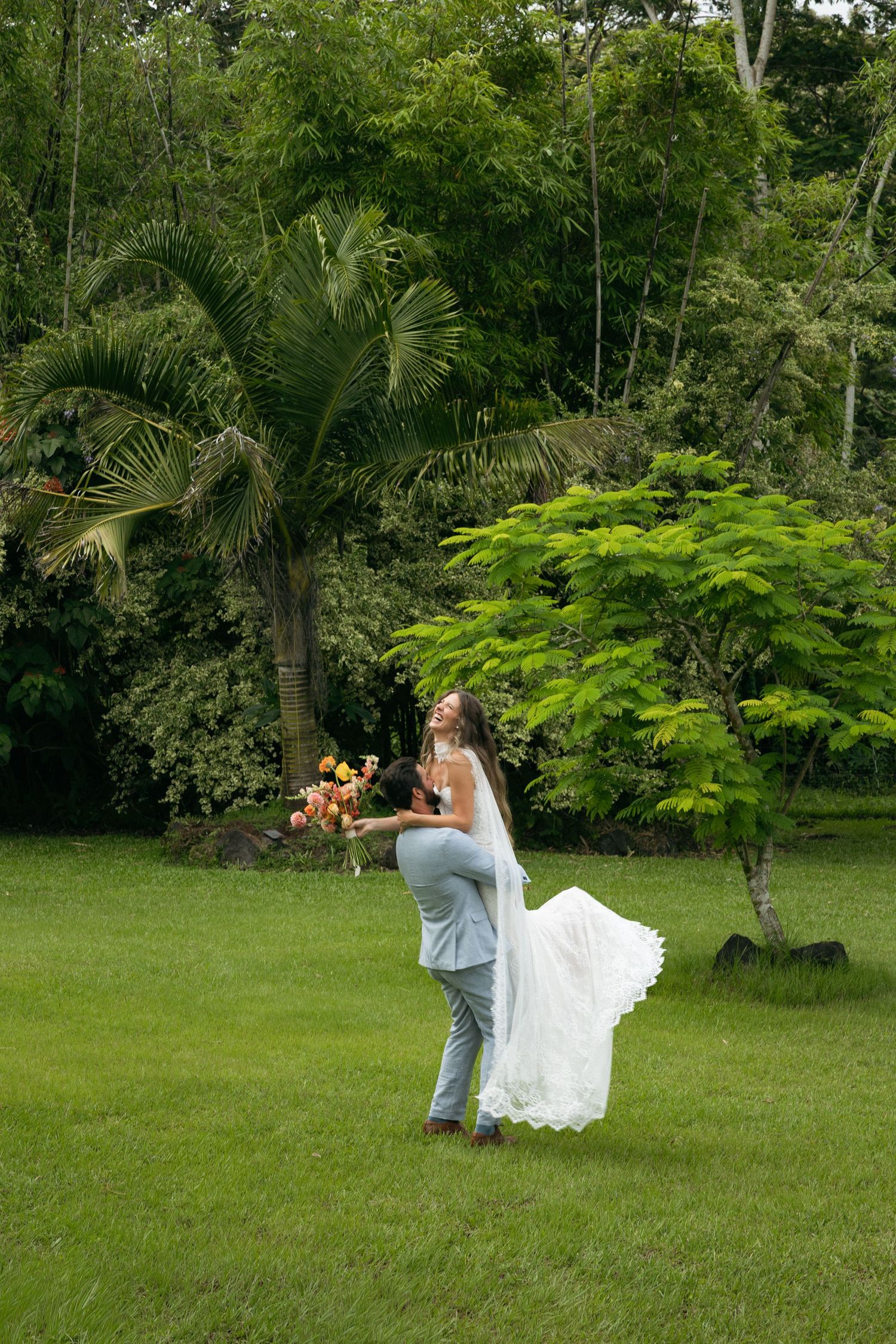 Groom lifting bride in white gown on lush green lawn surrounded by tropical trees during outdoor wedding celebration.
