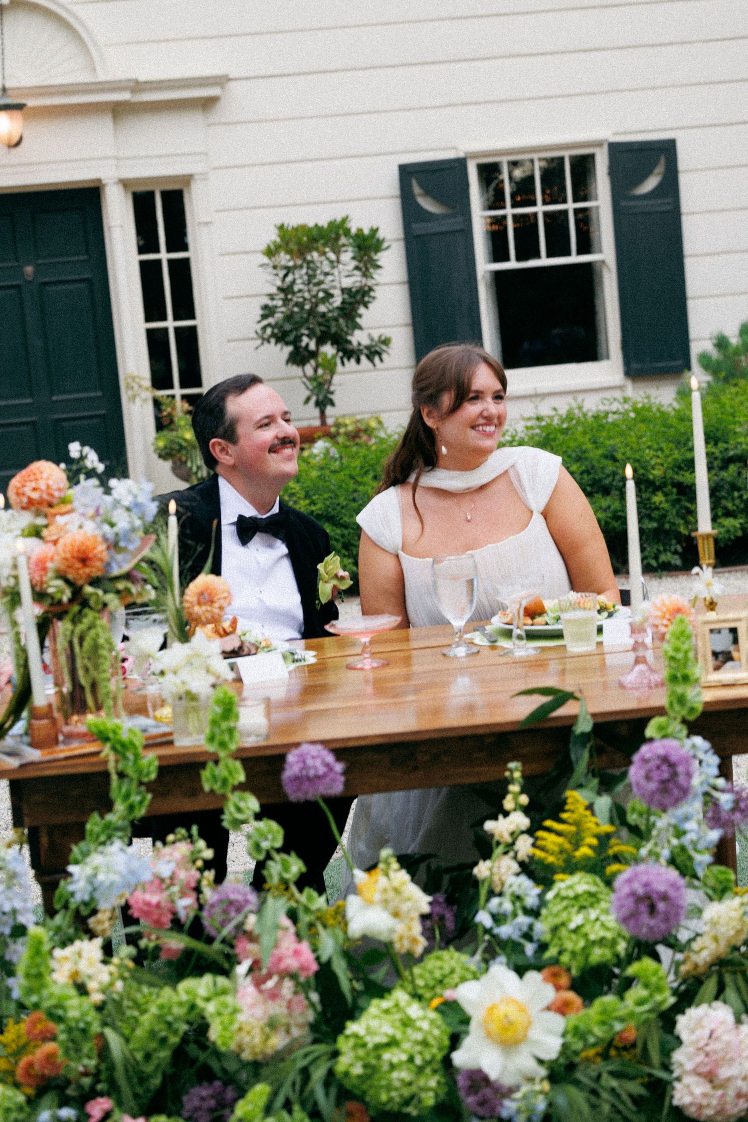 Bride and groom seated at outdoor reception table surrounded by lush colorful floral arrangements.