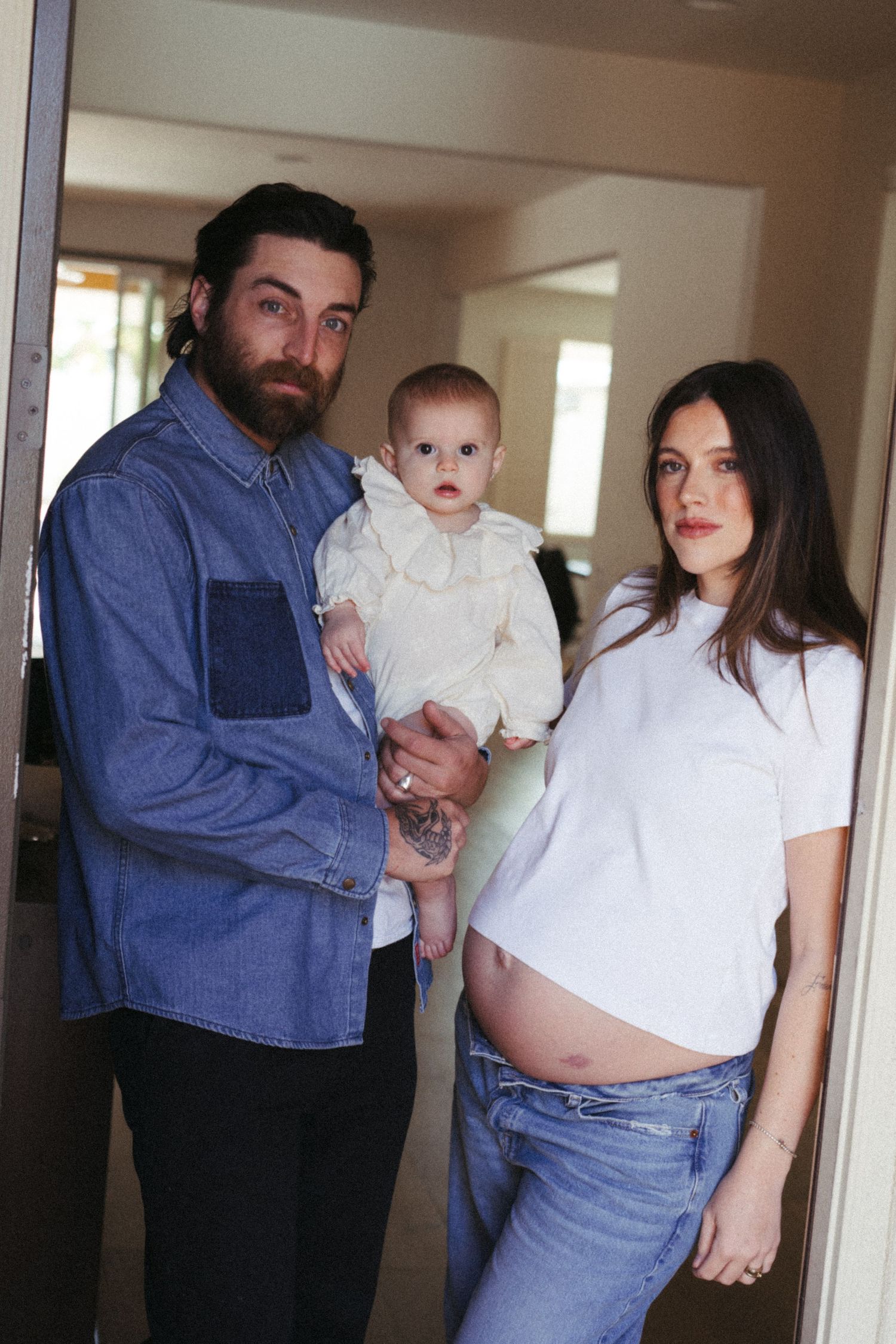 Pregnant woman in white t-shirt and jeans poses with bearded man holding baby in doorway.