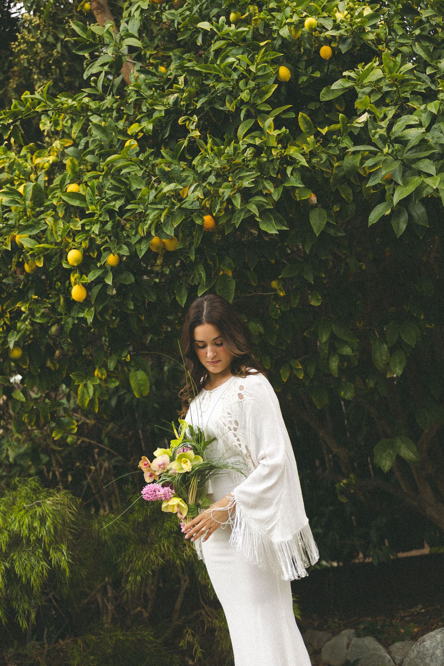 Bride in white fringe dress holding colorful bouquet standing beneath a lemon tree in a garden.