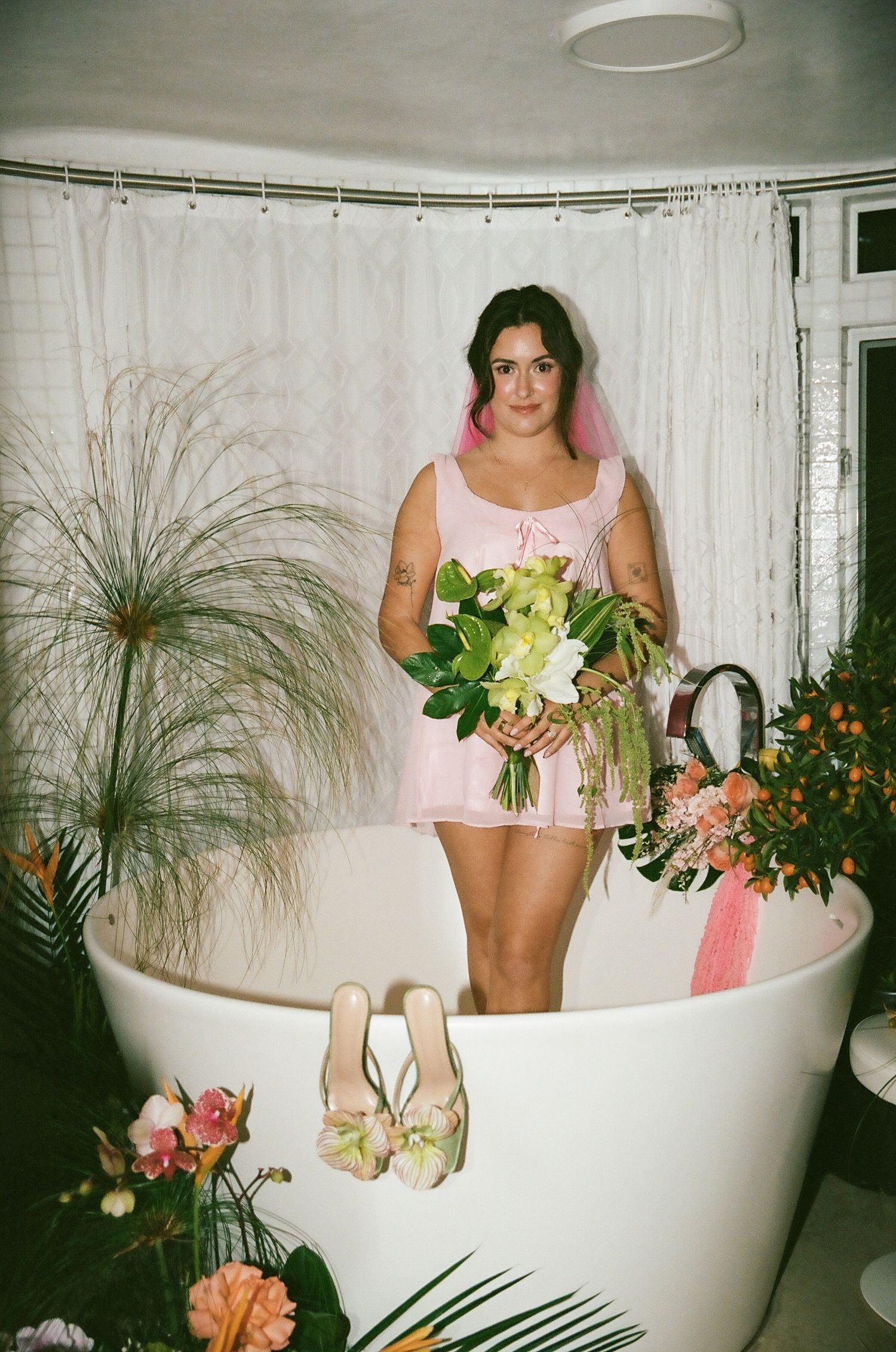 Bride in pink mini dress holding green bouquet standing in freestanding white bathtub with florals.