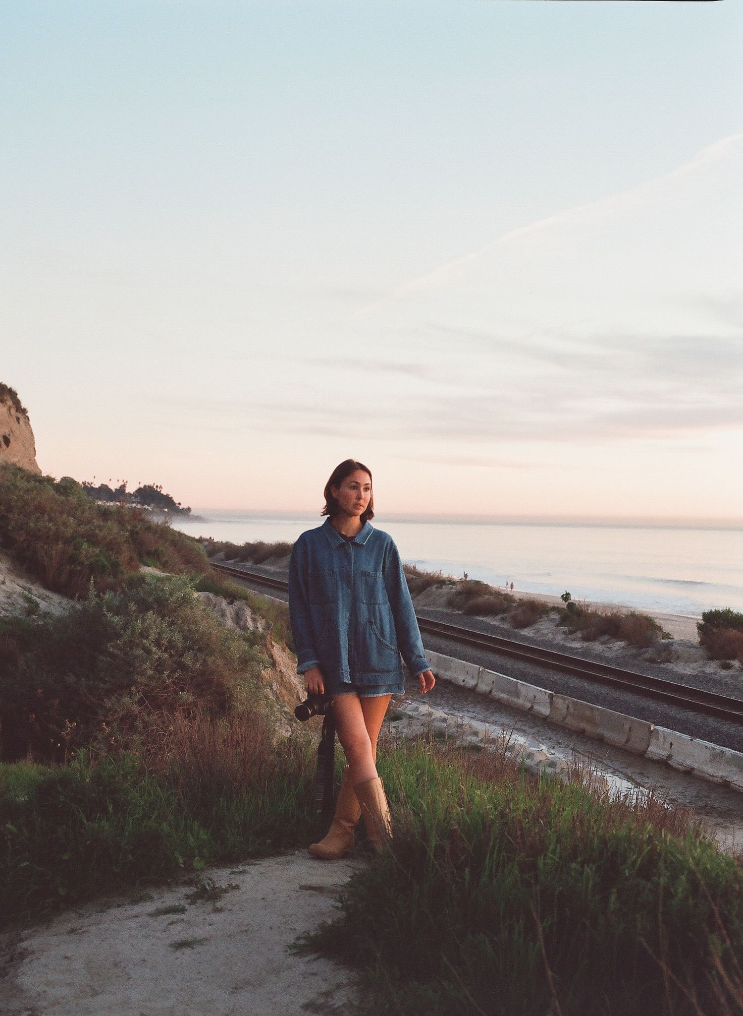 Young woman in denim jacket and shorts standing on coastal path near railway tracks at golden hour.