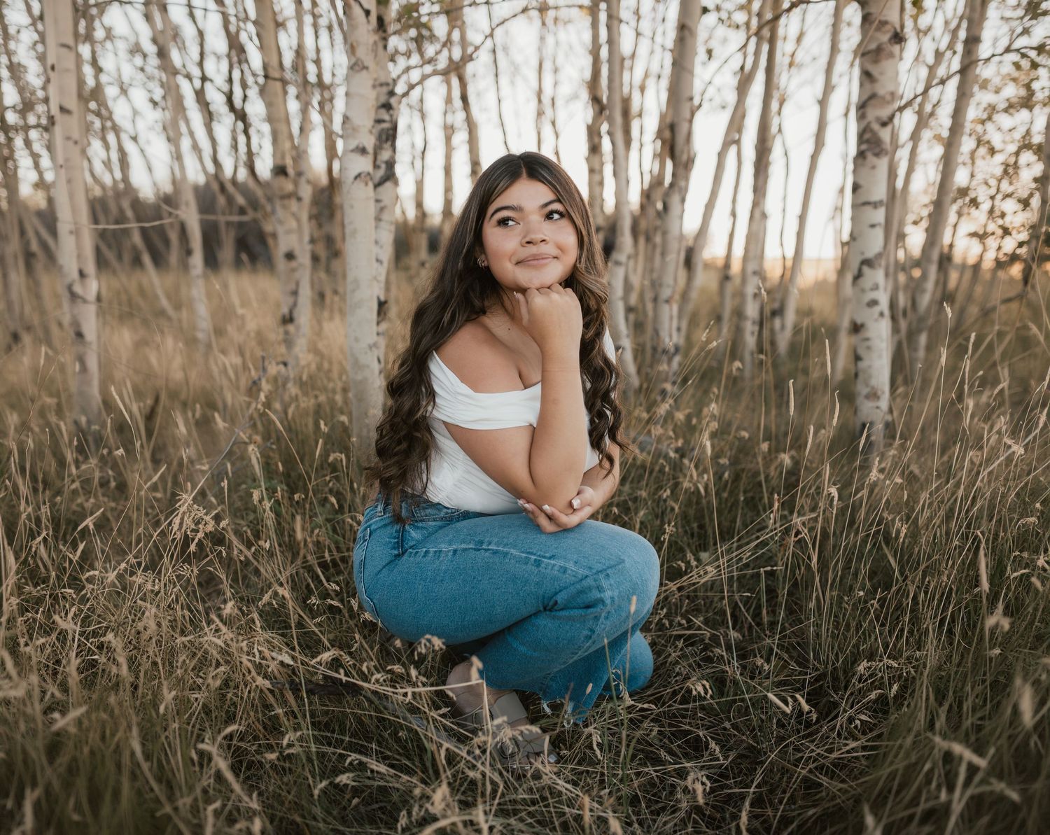 Young woman in white off-shoulder top and jeans sitting gracefully among birch trees in a golden field.