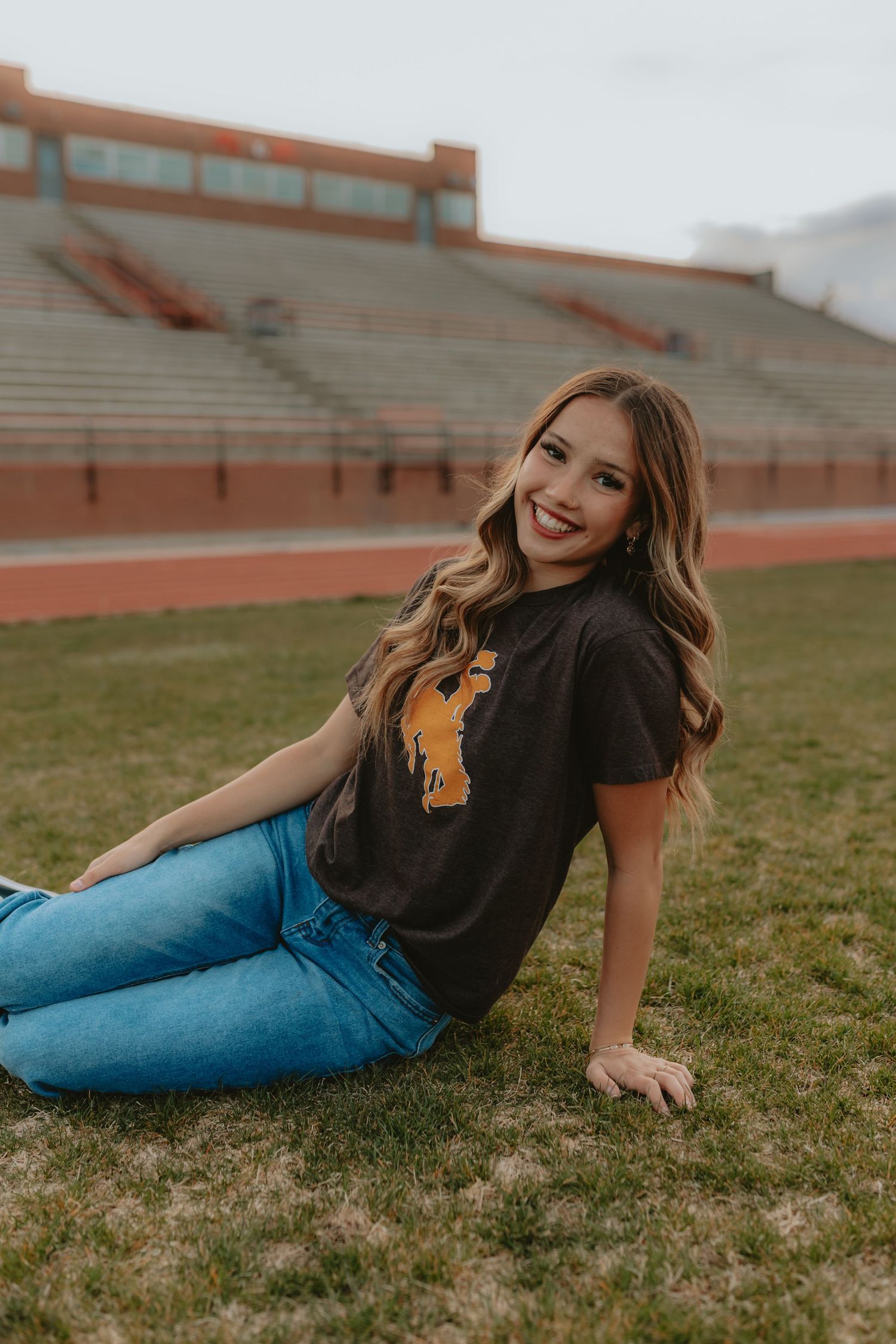 Young woman in dark graphic tee and jeans sitting on grass field near stadium bleachers, smiling.