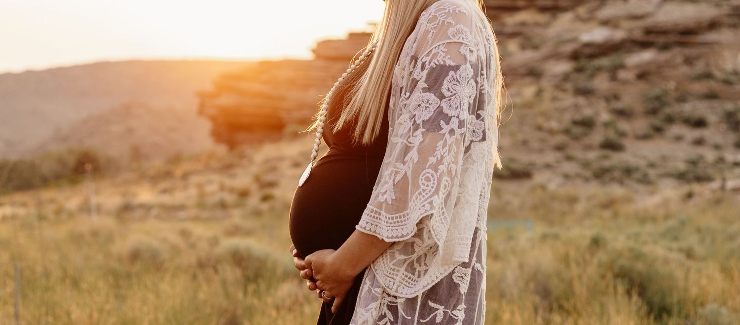A woman in a lace dress cradles her pregnant belly against a sunset backdrop in a field.
