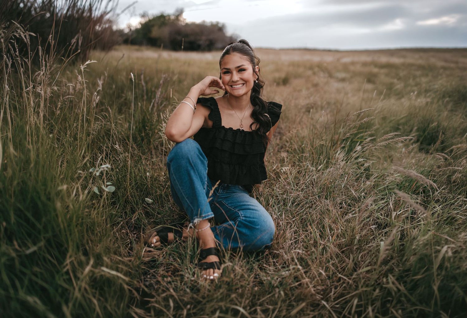 Young woman smiling in a field, wearing a black top and jeans, sitting in tall grass on a cloudy day.