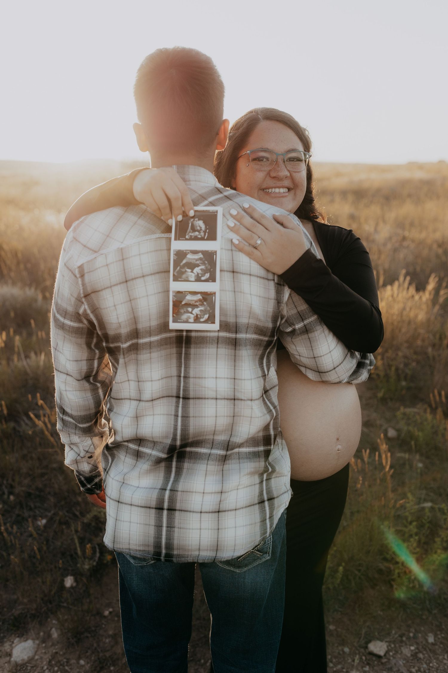 An expectant couple shares a tender moment while holding ultrasound photos in a golden field at sunset.