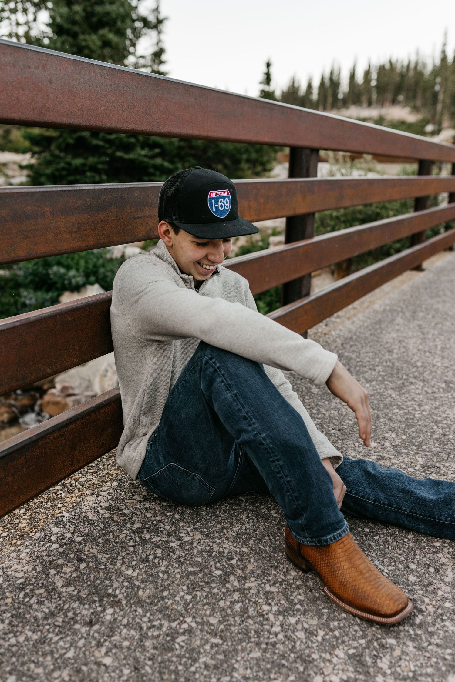 Man sitting on bridge wearing black cap, cream sweater, jeans, and brown cowboy boots, smiling outdoors.