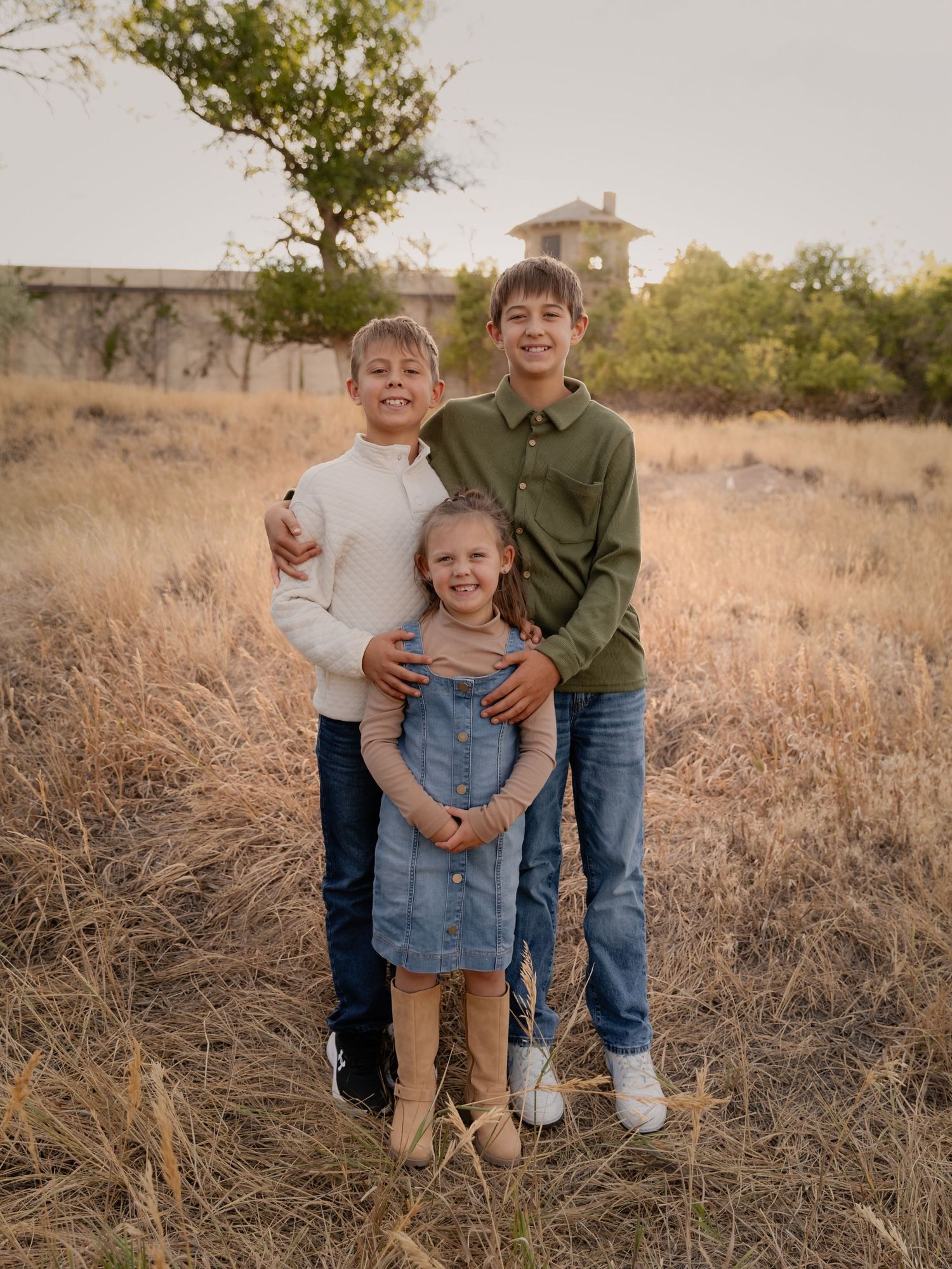Three children pose together in a golden field at sunset, two older boys flanking a young girl in a denim dress.