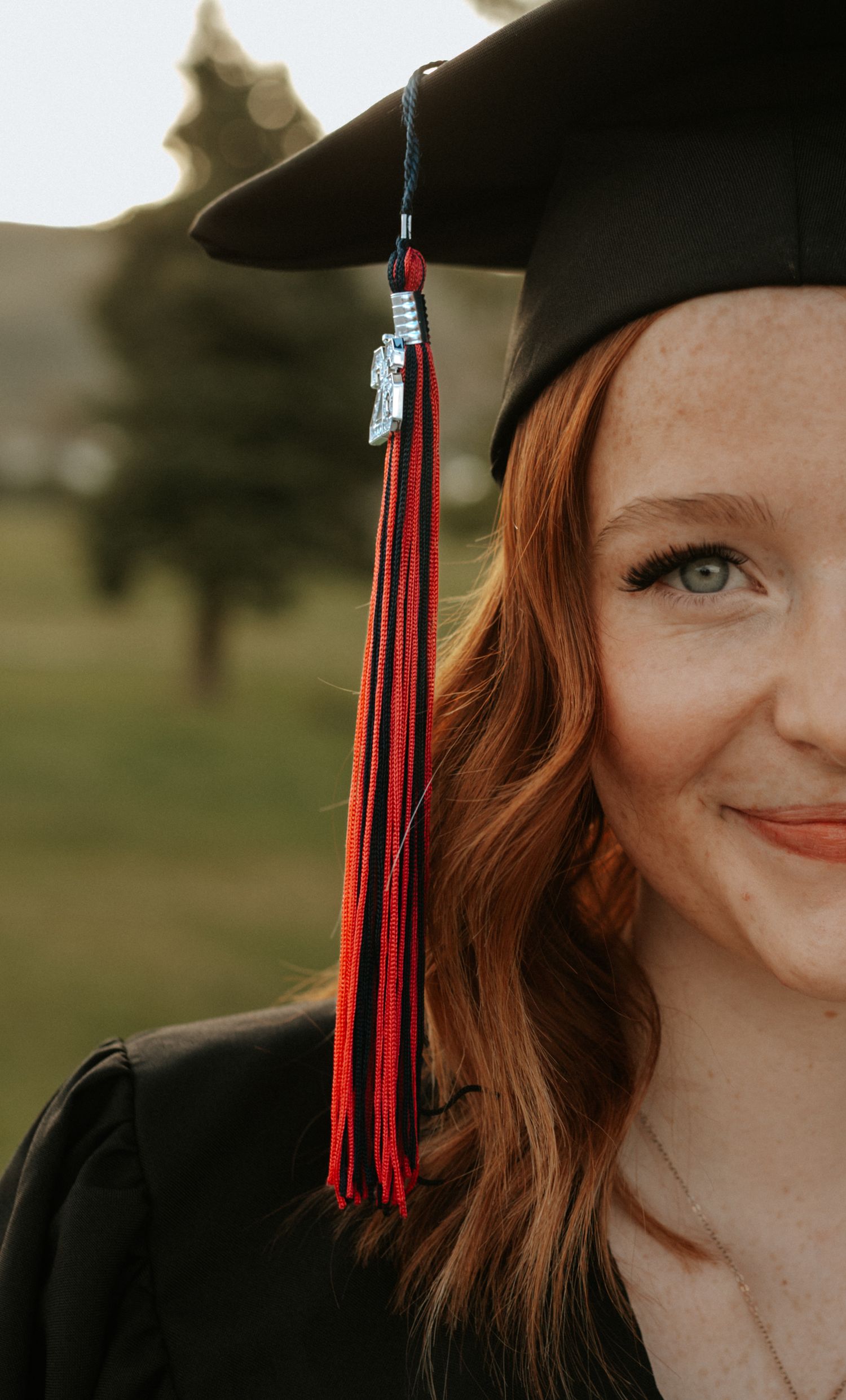 Young woman with red hair smiling in black graduation cap and gown with red tassel outdoors.