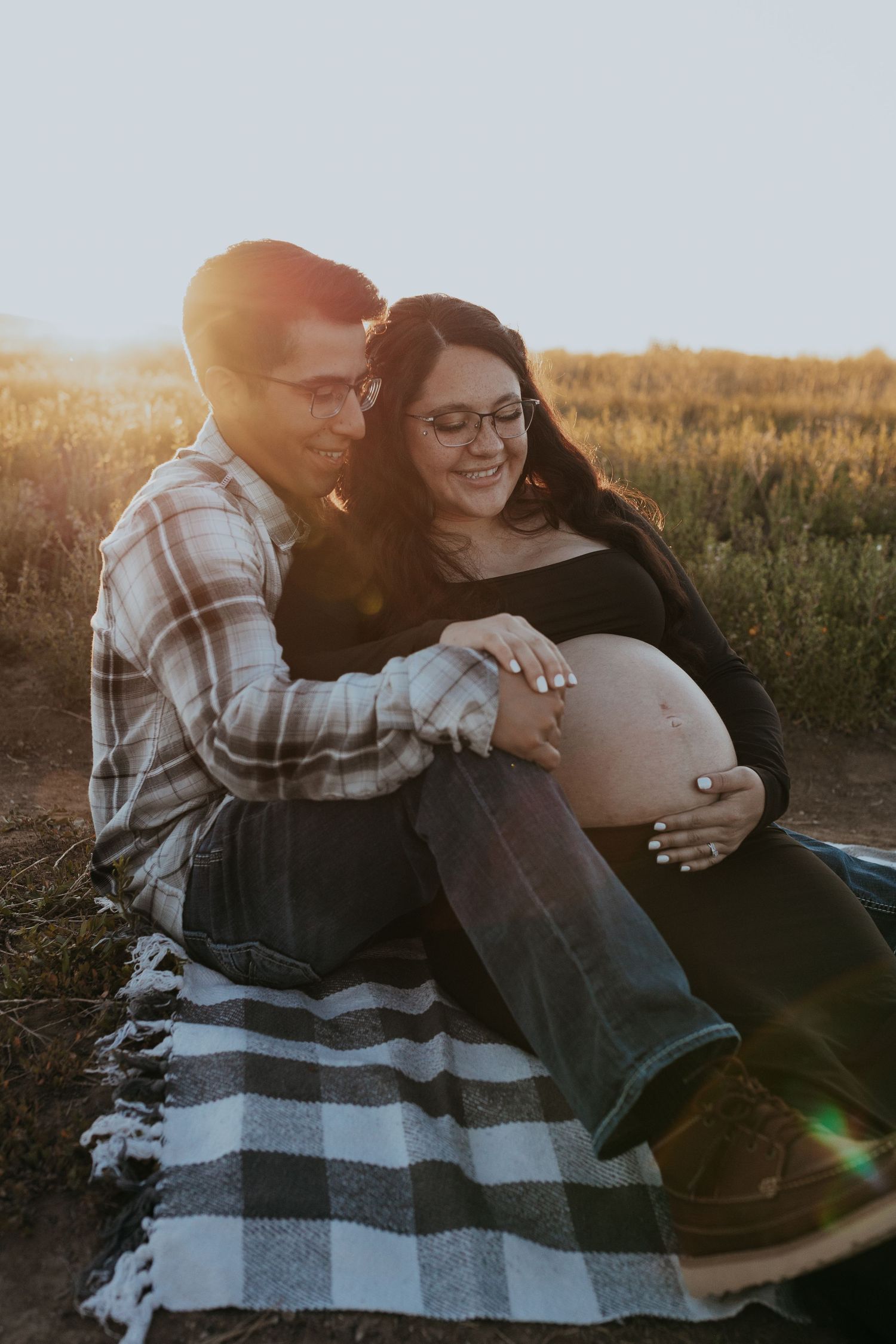 A couple sits on a plaid blanket in a field during their maternity photo session at golden hour.