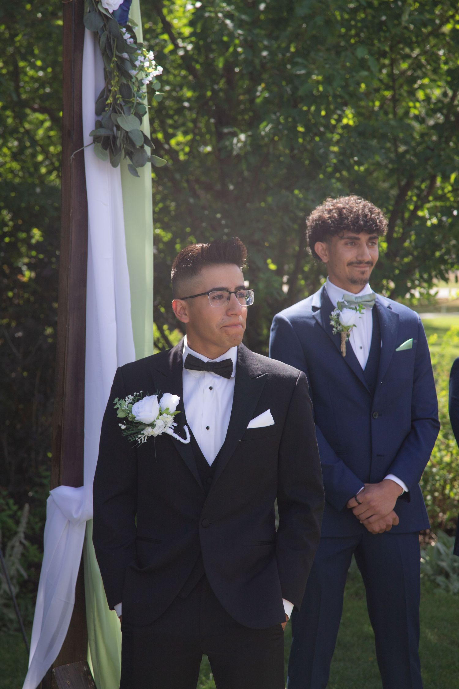 Two formally dressed people stand at a wedding ceremony arch decorated with white fabric and greenery outdoors.