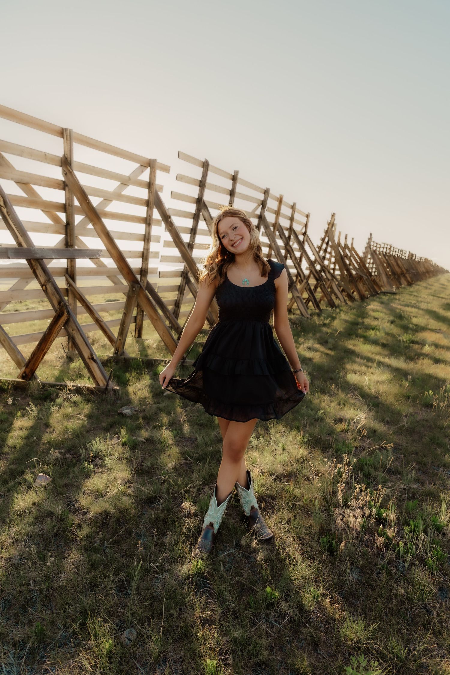 Young woman in black dress and cowboy boots smiling beside a wooden fence on a sunny day.