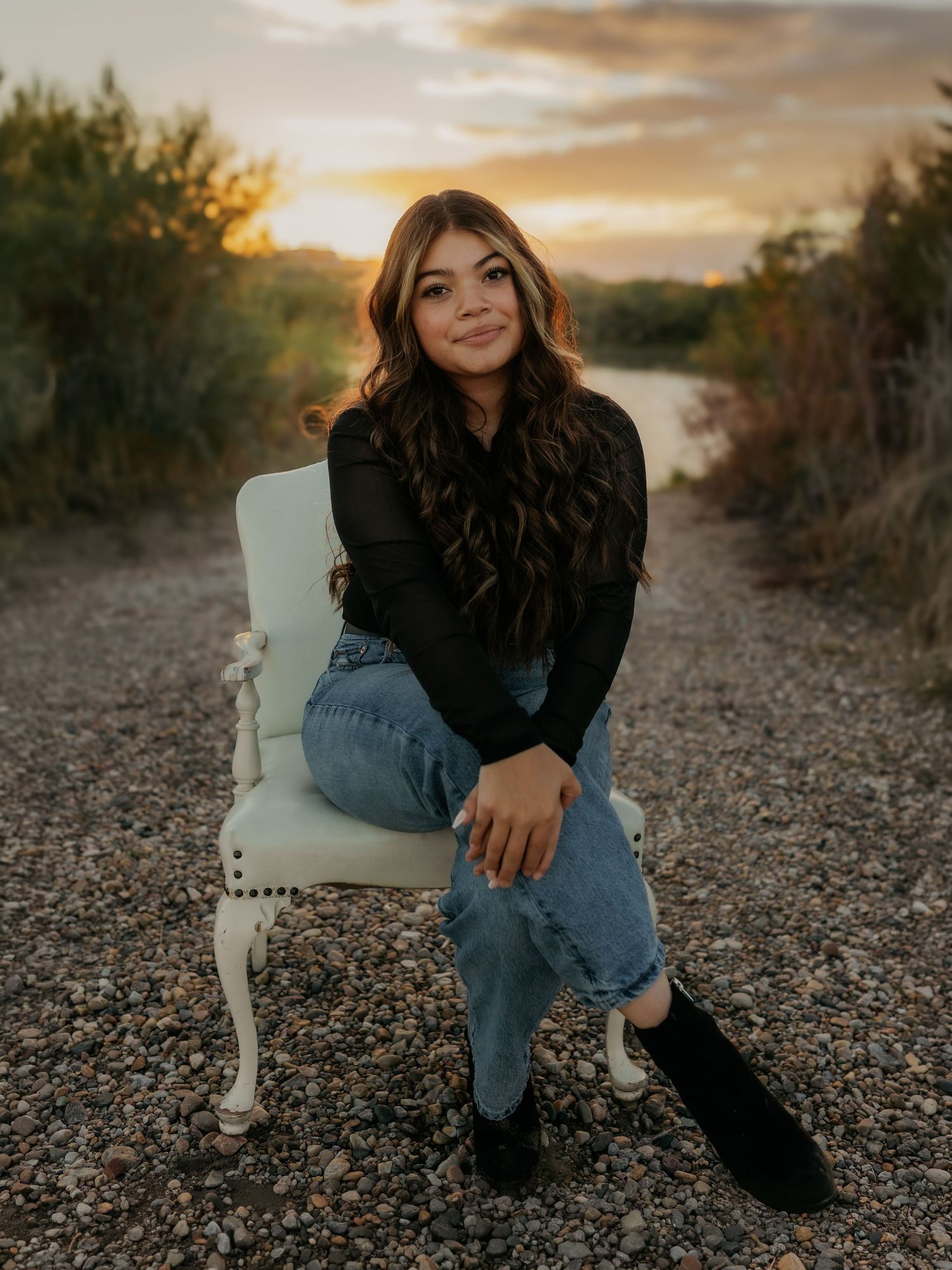 Young woman in black top and jeans sits on white chair on gravel path at golden hour sunset.