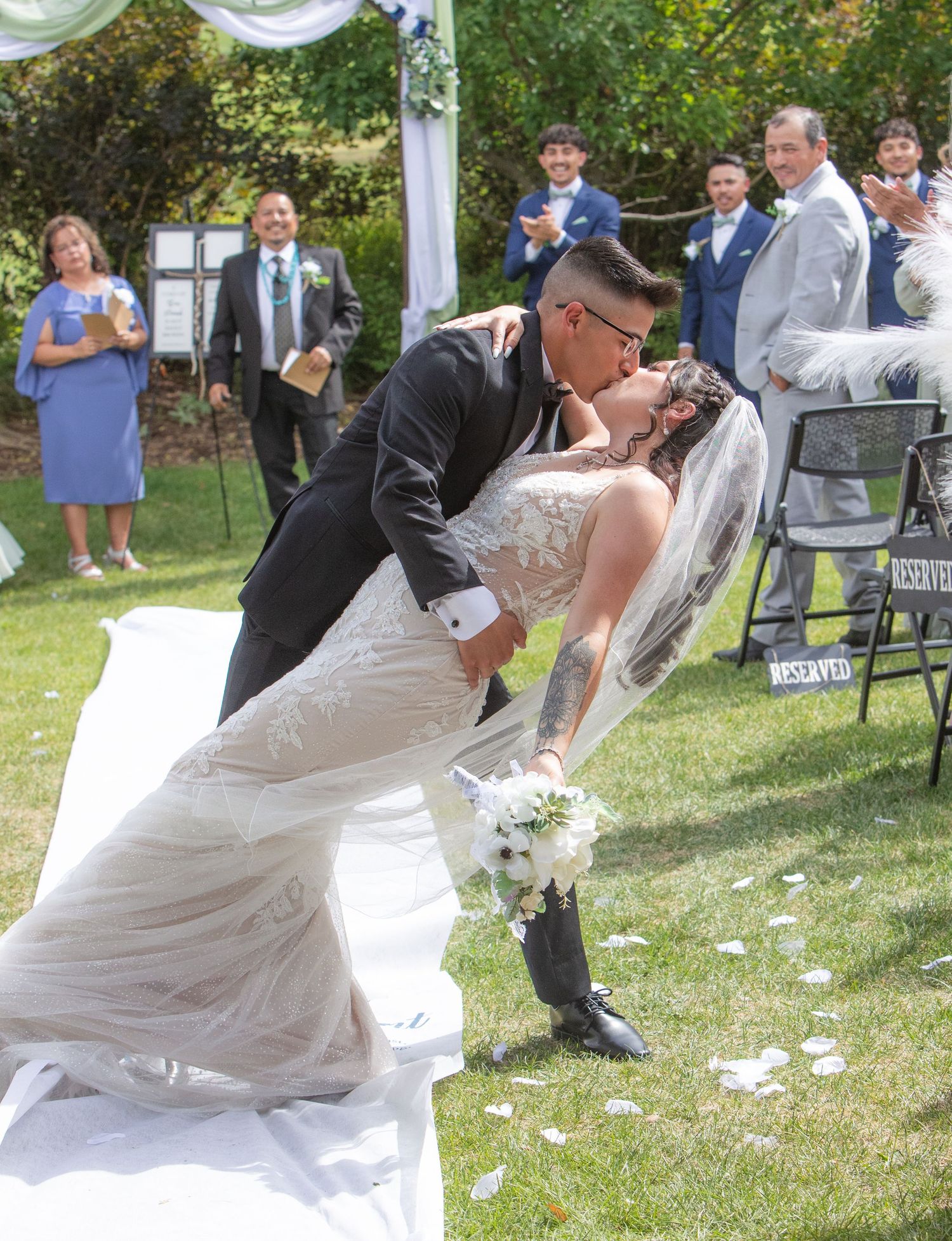 Newlyweds share a romantic dip kiss during outdoor garden wedding ceremony with guests watching in background.