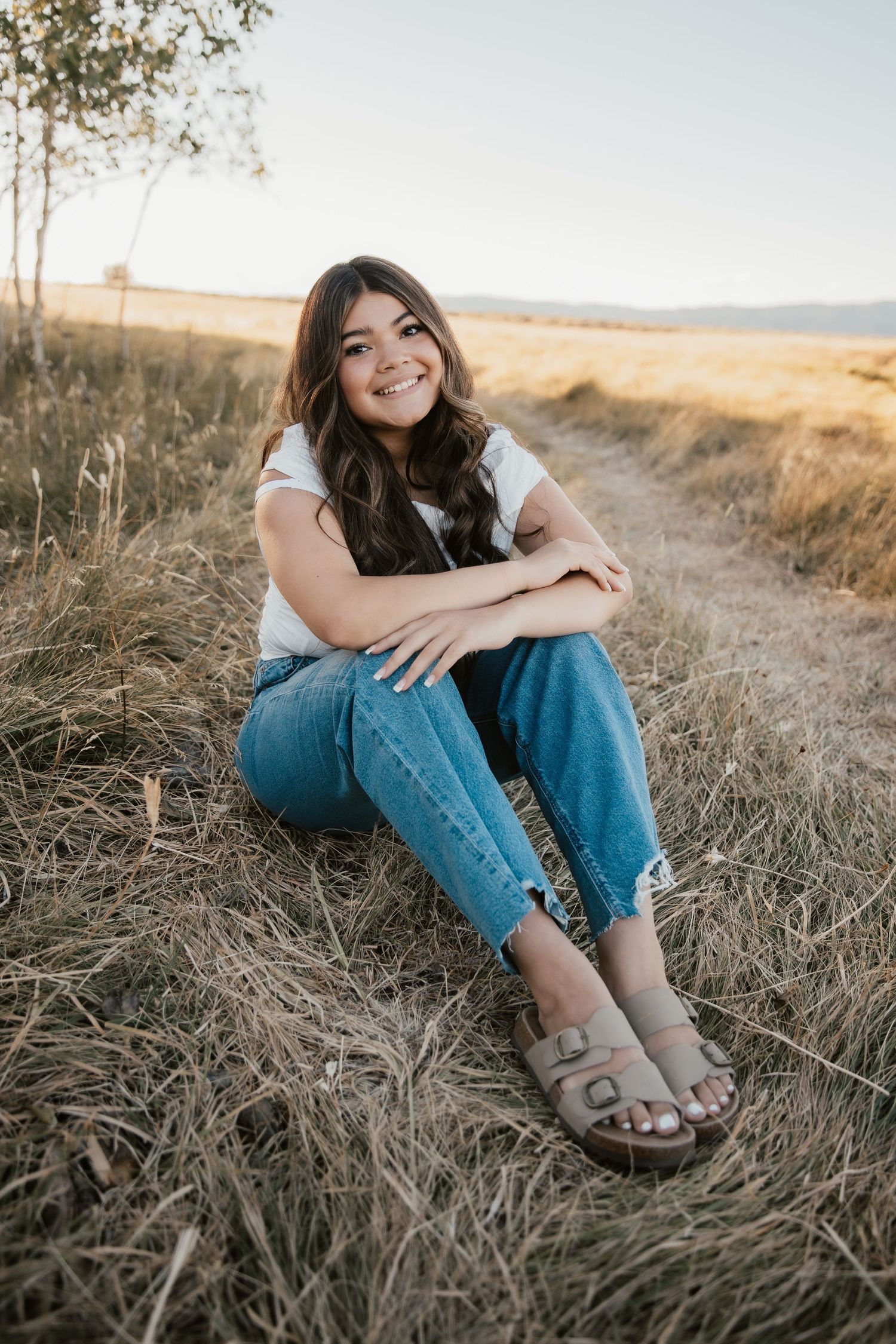 Young woman with long dark hair sits smiling in a golden field, wearing white tee, jeans, and sandals.