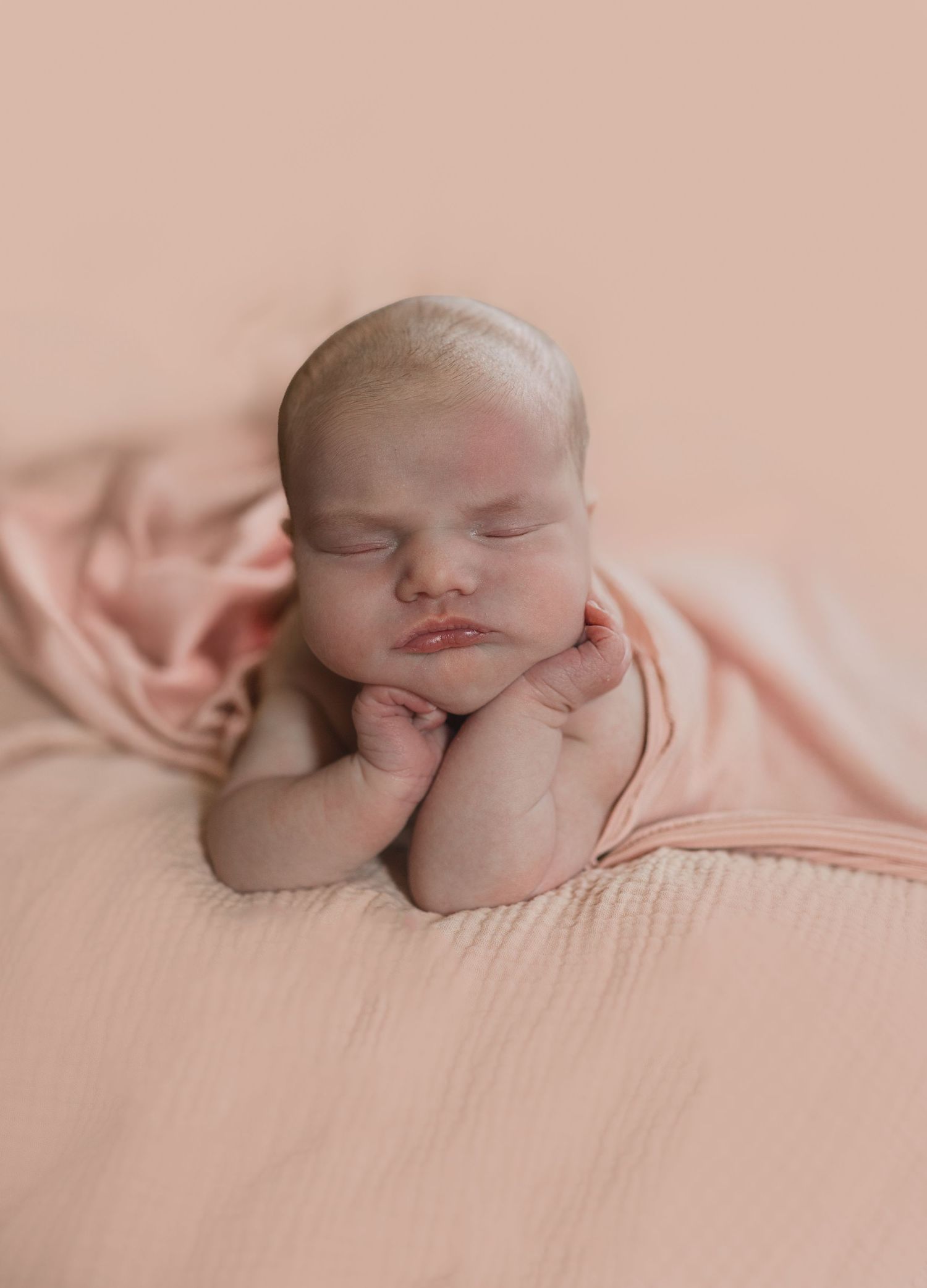 Sleeping newborn posed on pink fabric with hands under chin.