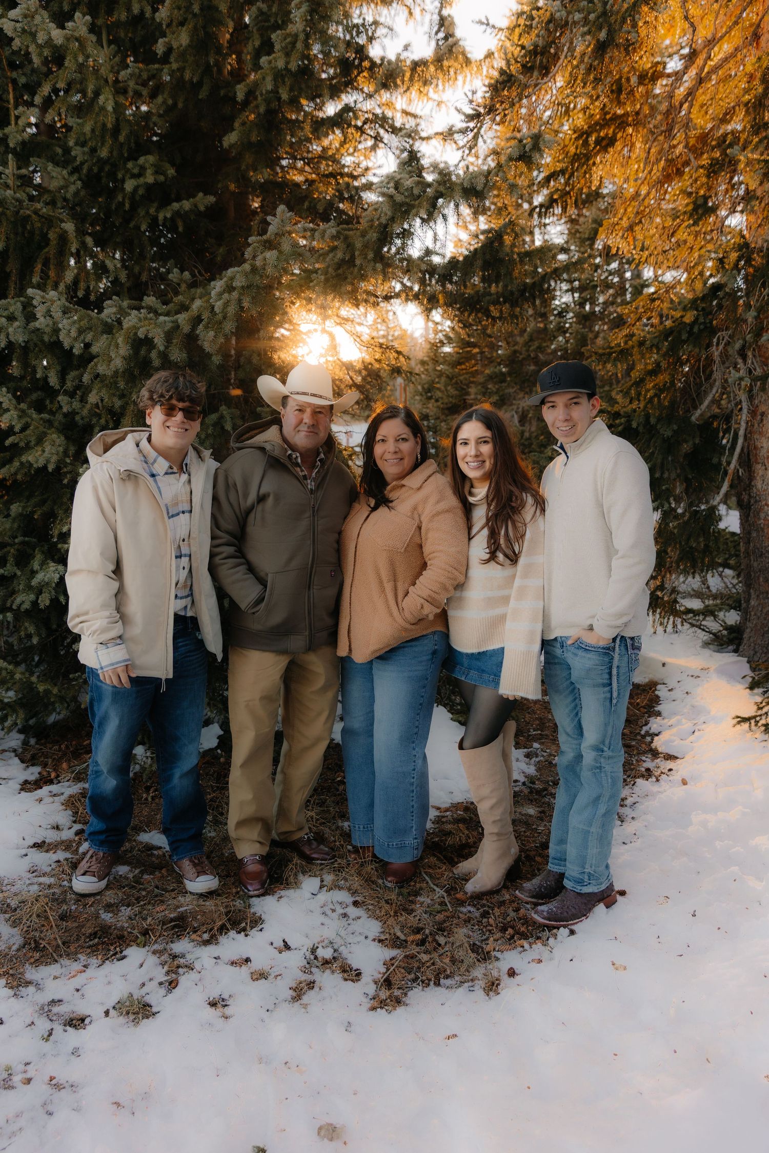 Family of five posing outdoors in winter snow among evergreen trees at golden sunset hour.