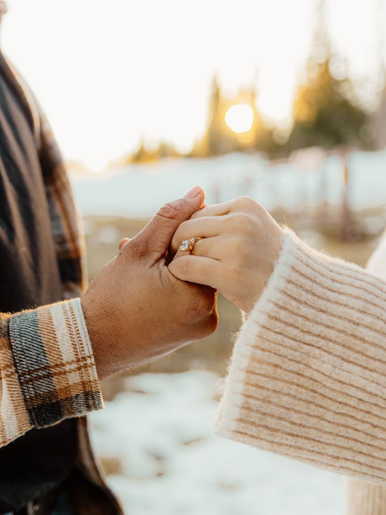 Two hands holding each other with engagement ring visible against warm sunset lighting in winter setting.