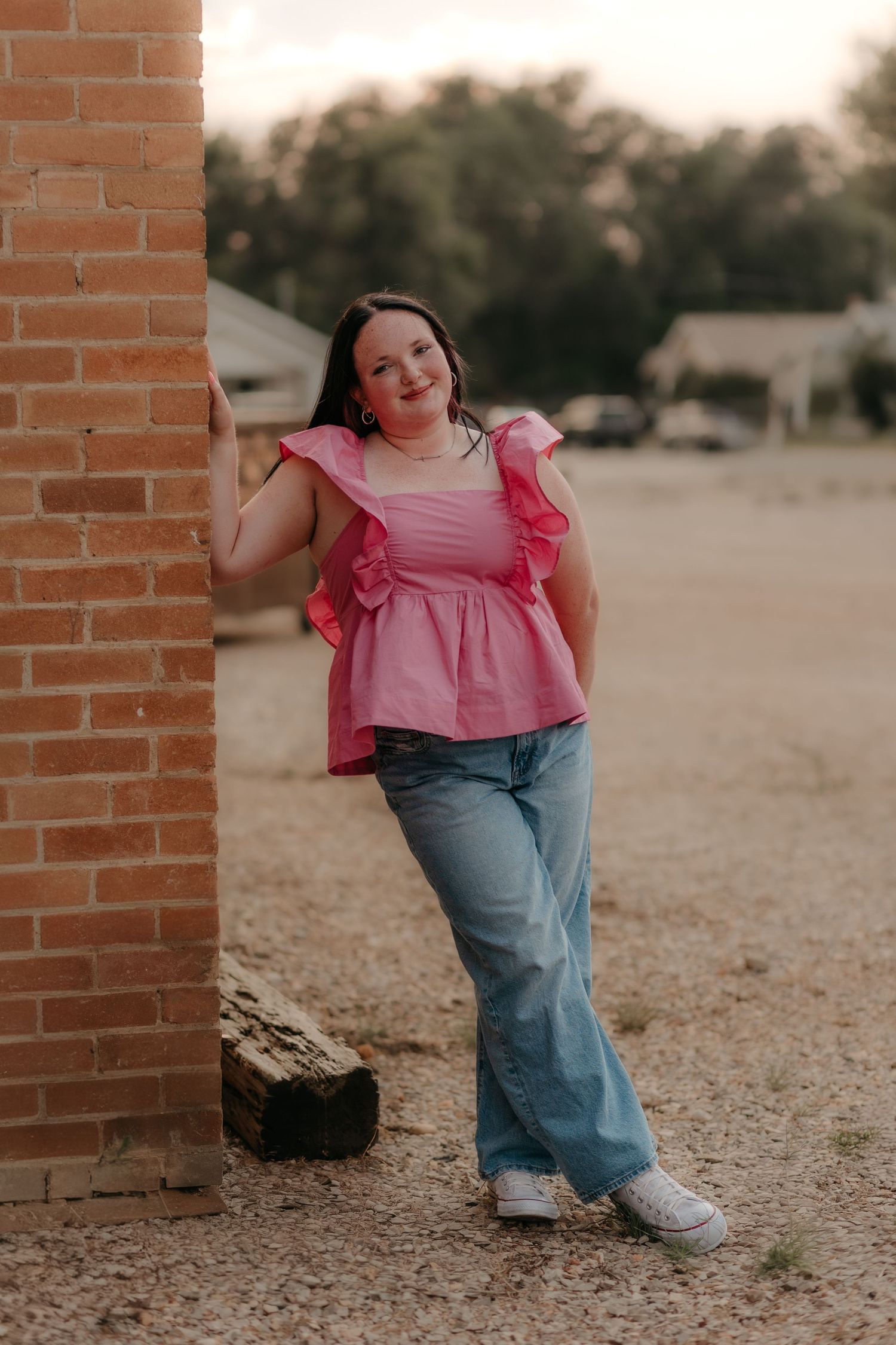 Young woman in pink ruffled top and flared jeans leans against brick wall outdoors, smiling confidently.