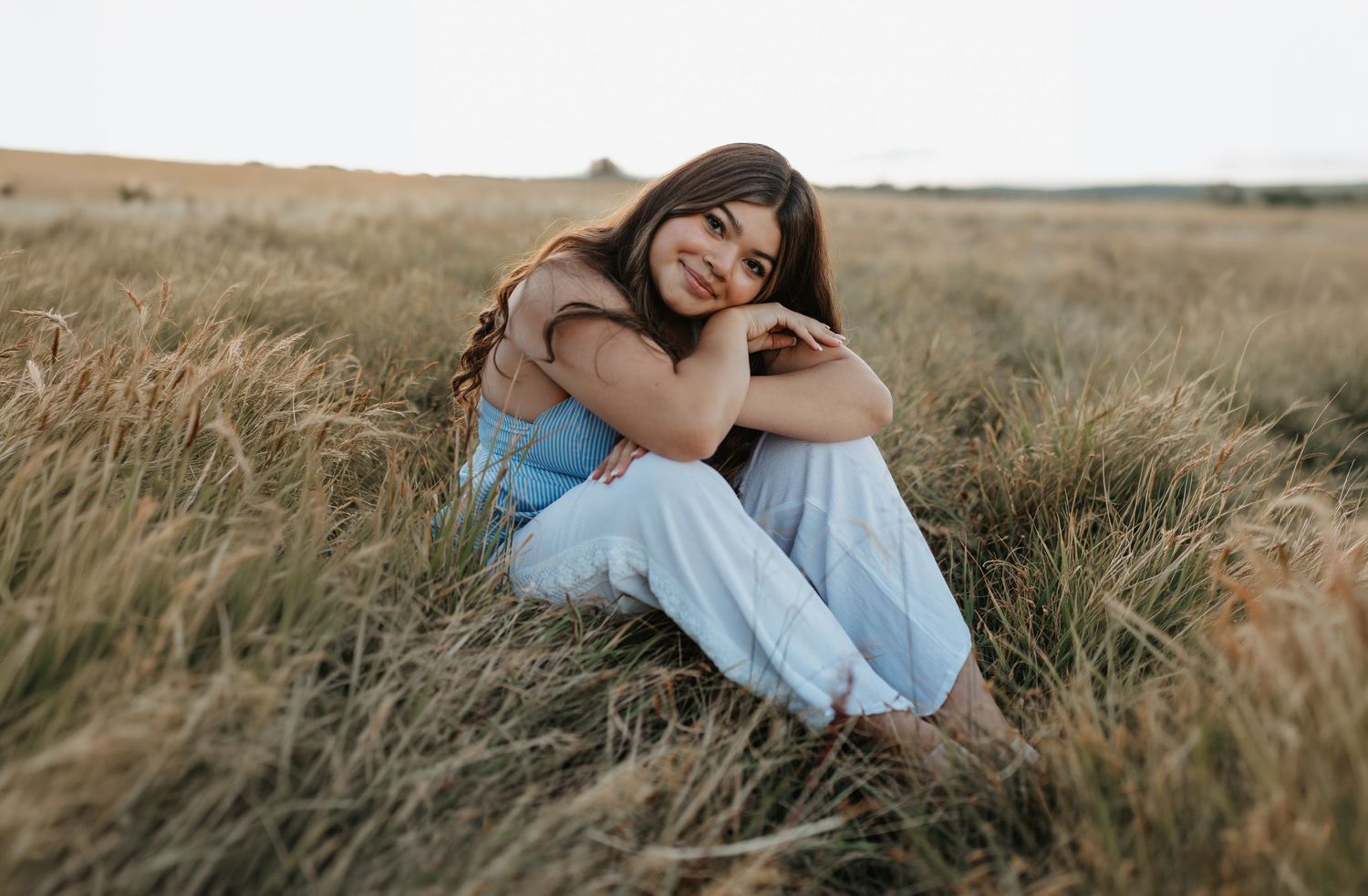 Young woman with brown hair sitting in a golden grass field, smiling warmly in a light blue outfit.