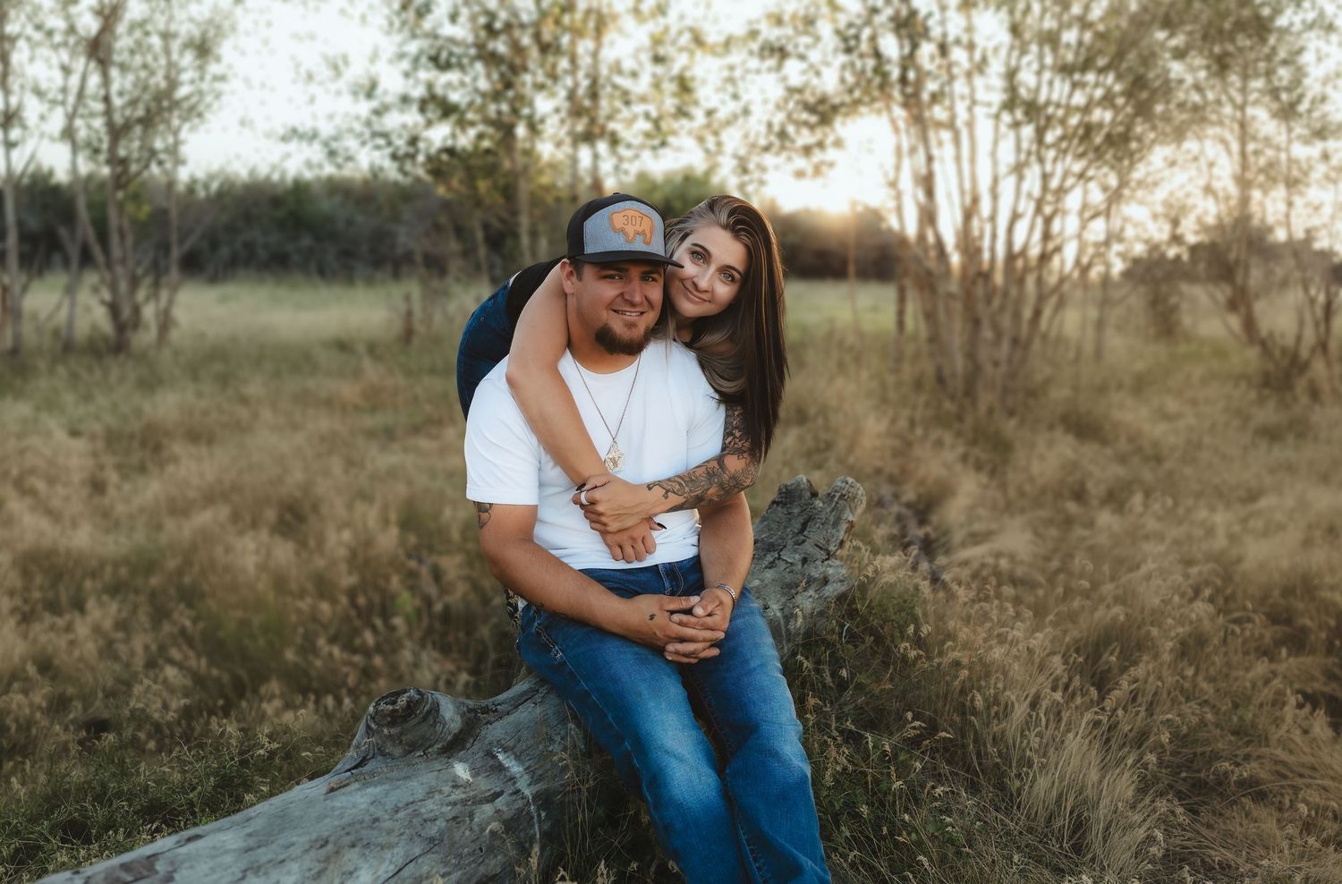 Couple embracing on a fallen log in a golden field at sunset, man in white tee and cap, woman in jeans smiling.