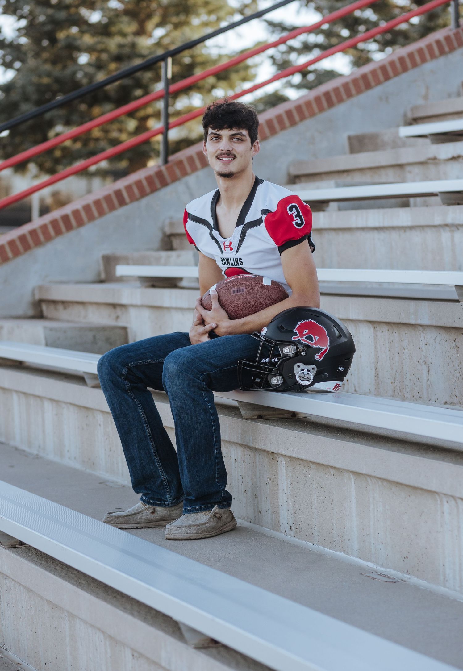 Football player in white jersey sitting on stadium steps with helmet and ball.