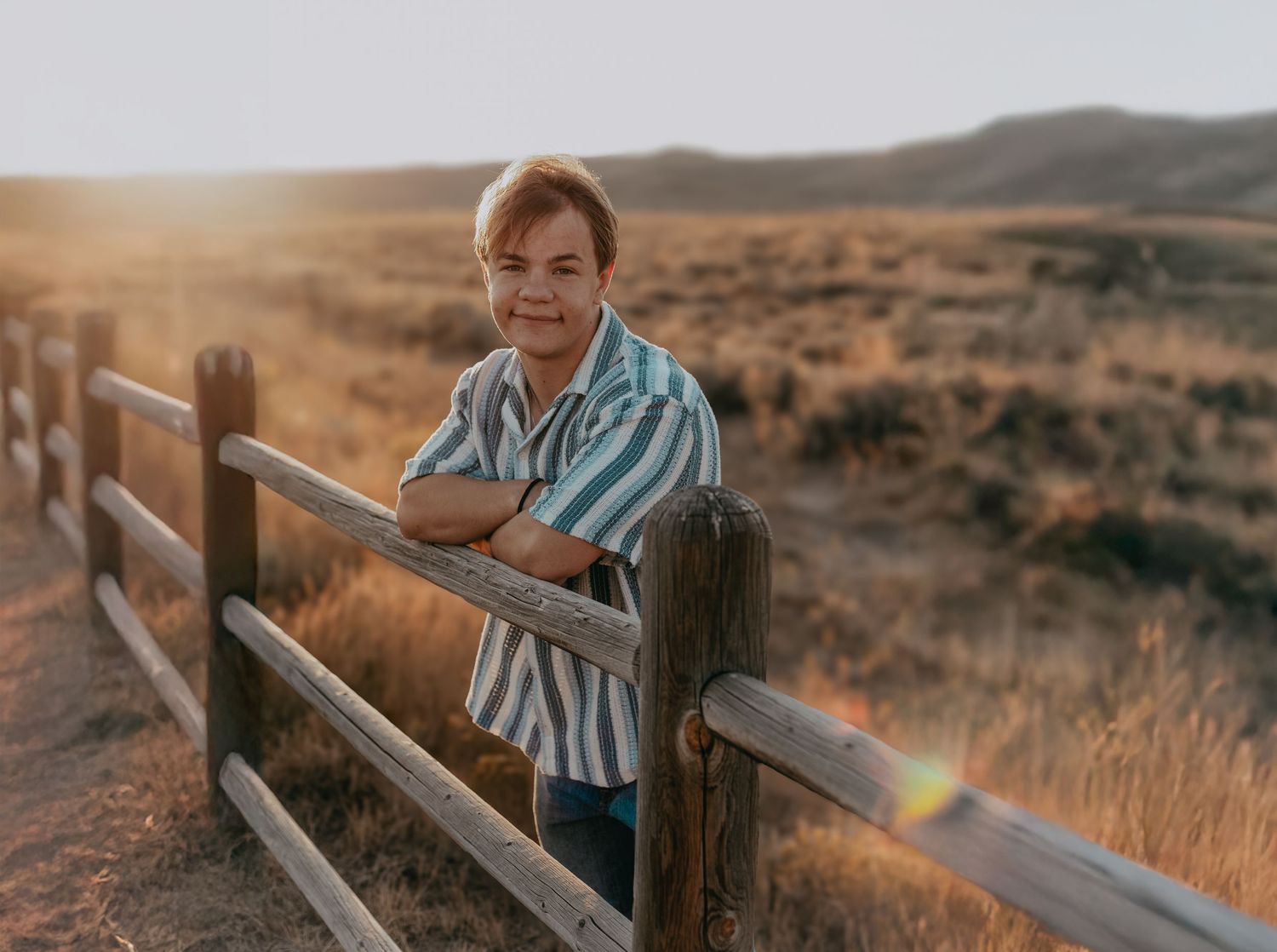 Young man leaning on a wooden fence at golden hour with scenic hills in the background.