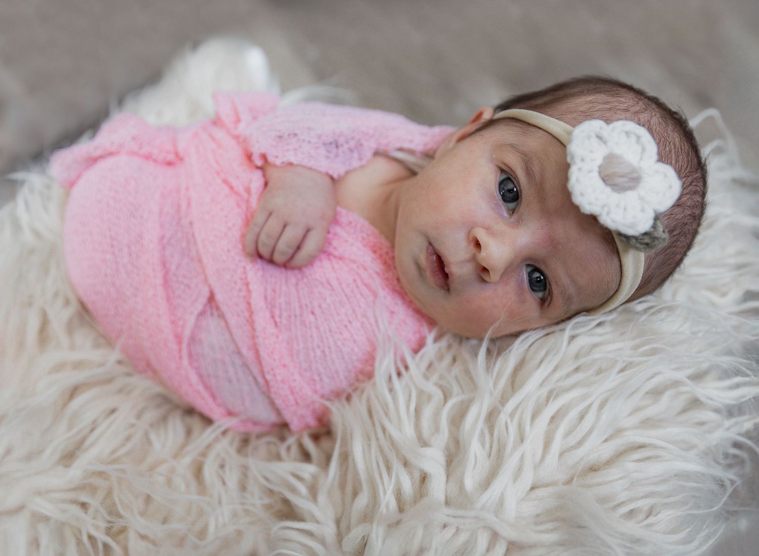 Baby in pink wrap with floral headband looking at camera on white furry backdrop.