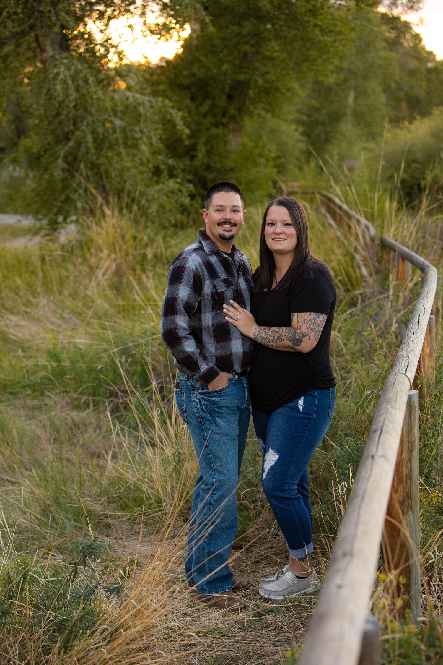 Couple standing by wooden railing at sunset in rustic outdoor setting.