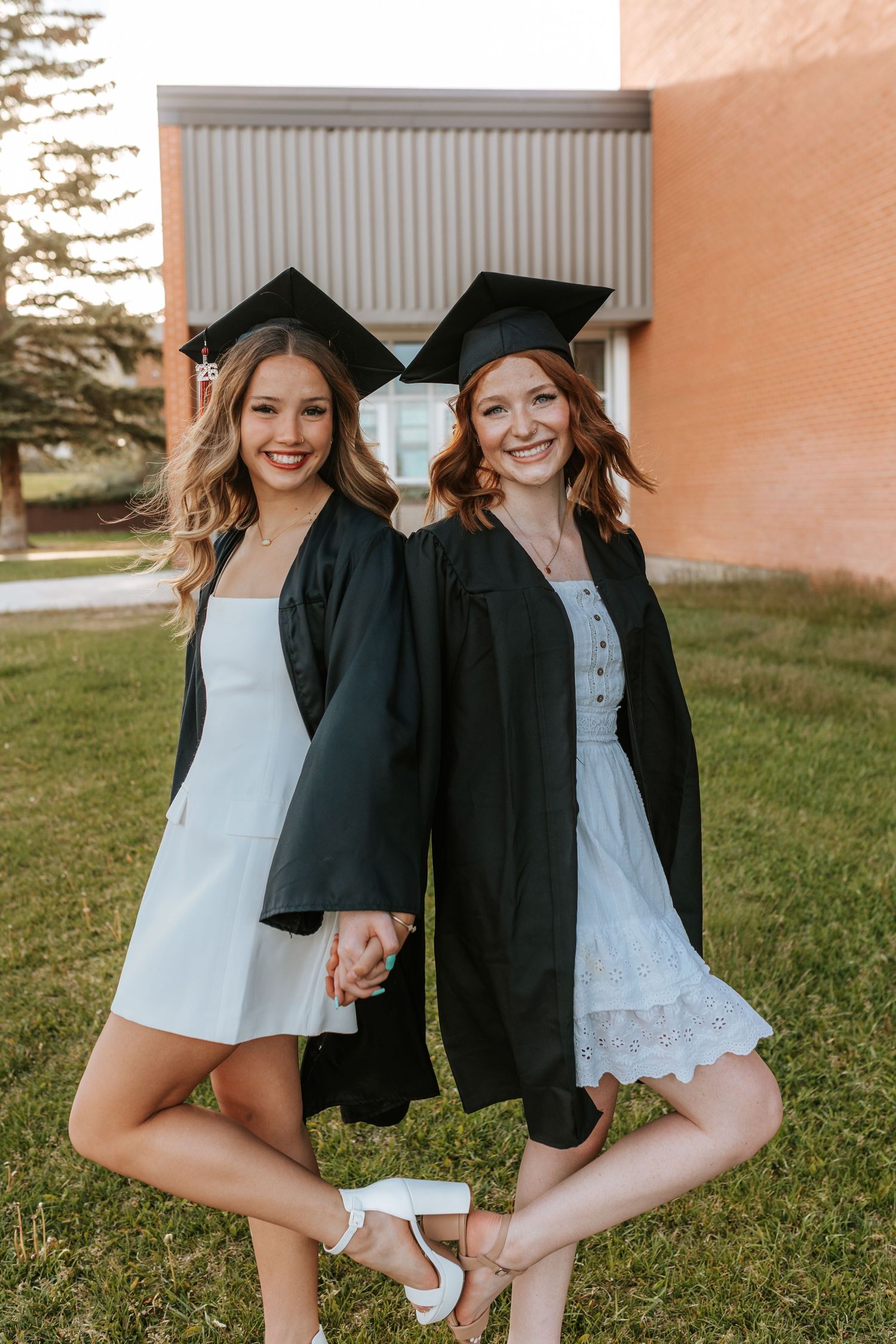 Two smiling women in black graduation caps and gowns over white dresses pose outdoors on a sunny day.